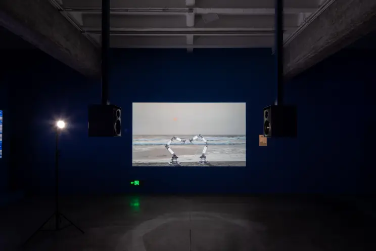 Gallery display with a screen showing mechanical arms on a beach between speakers.