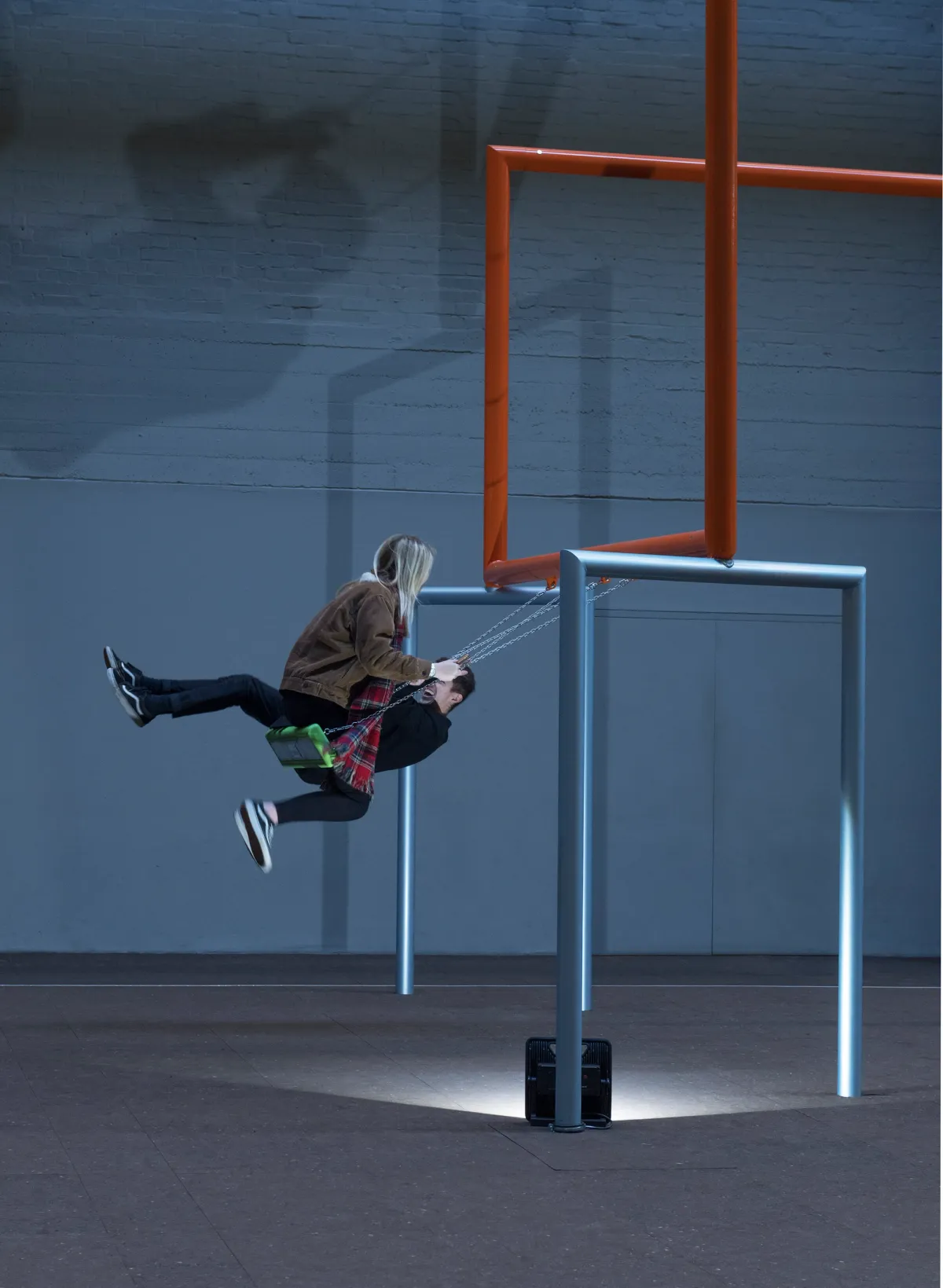 Two individuals swinging on swings suspended from orange and grey pipes in a spacious art gallery.