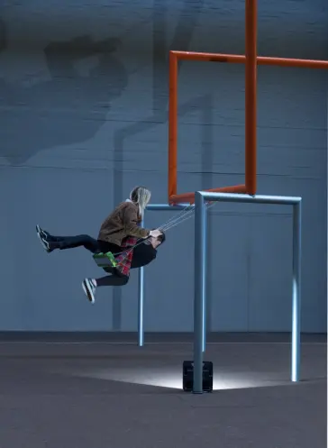 Two individuals swinging on swings suspended from orange and grey pipes in a spacious art gallery.