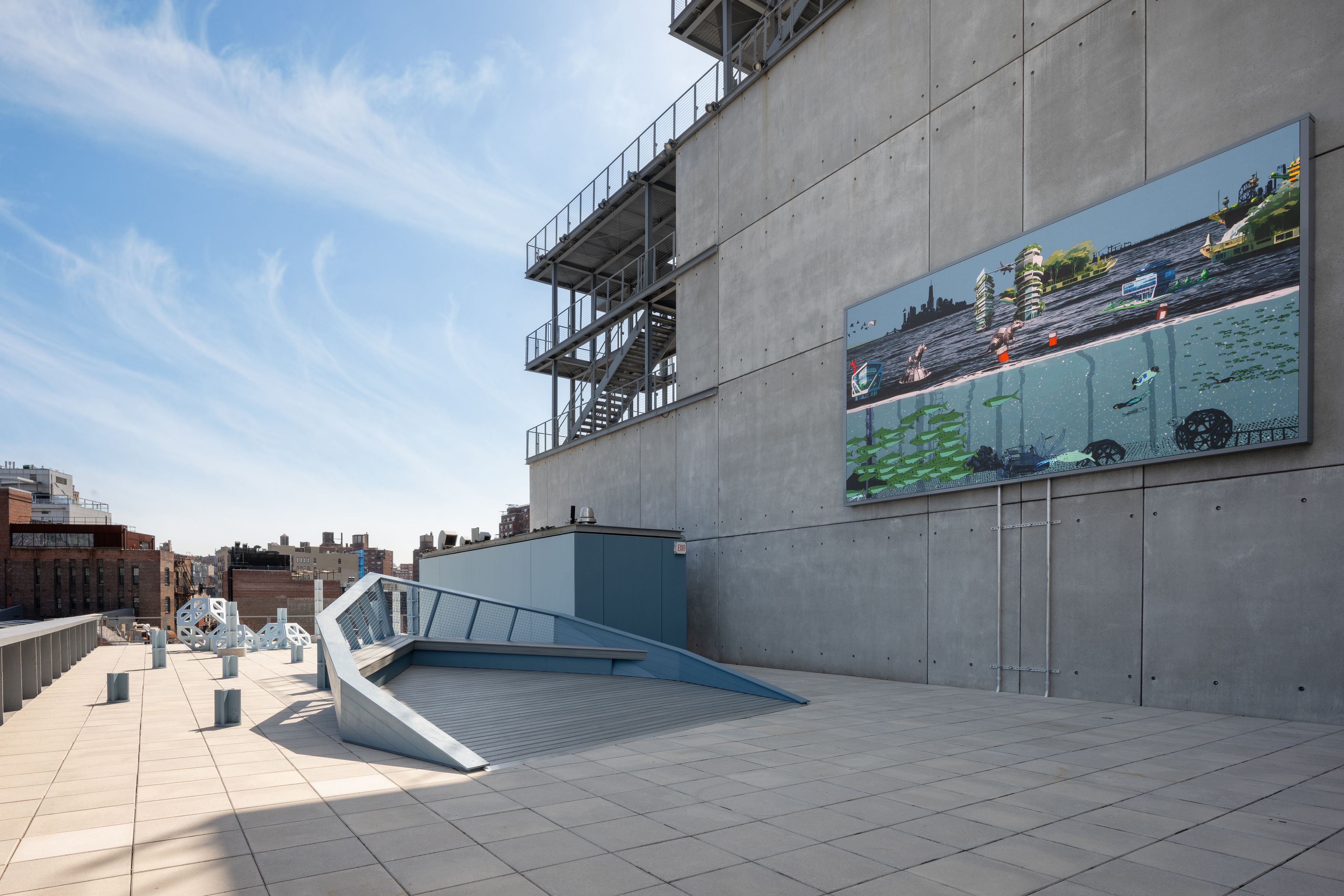 A blue sky on the left of the photo faces a building rooftop. Several blue sculptures, one in the form of a ship, take up space. An LED screen plays an animation.