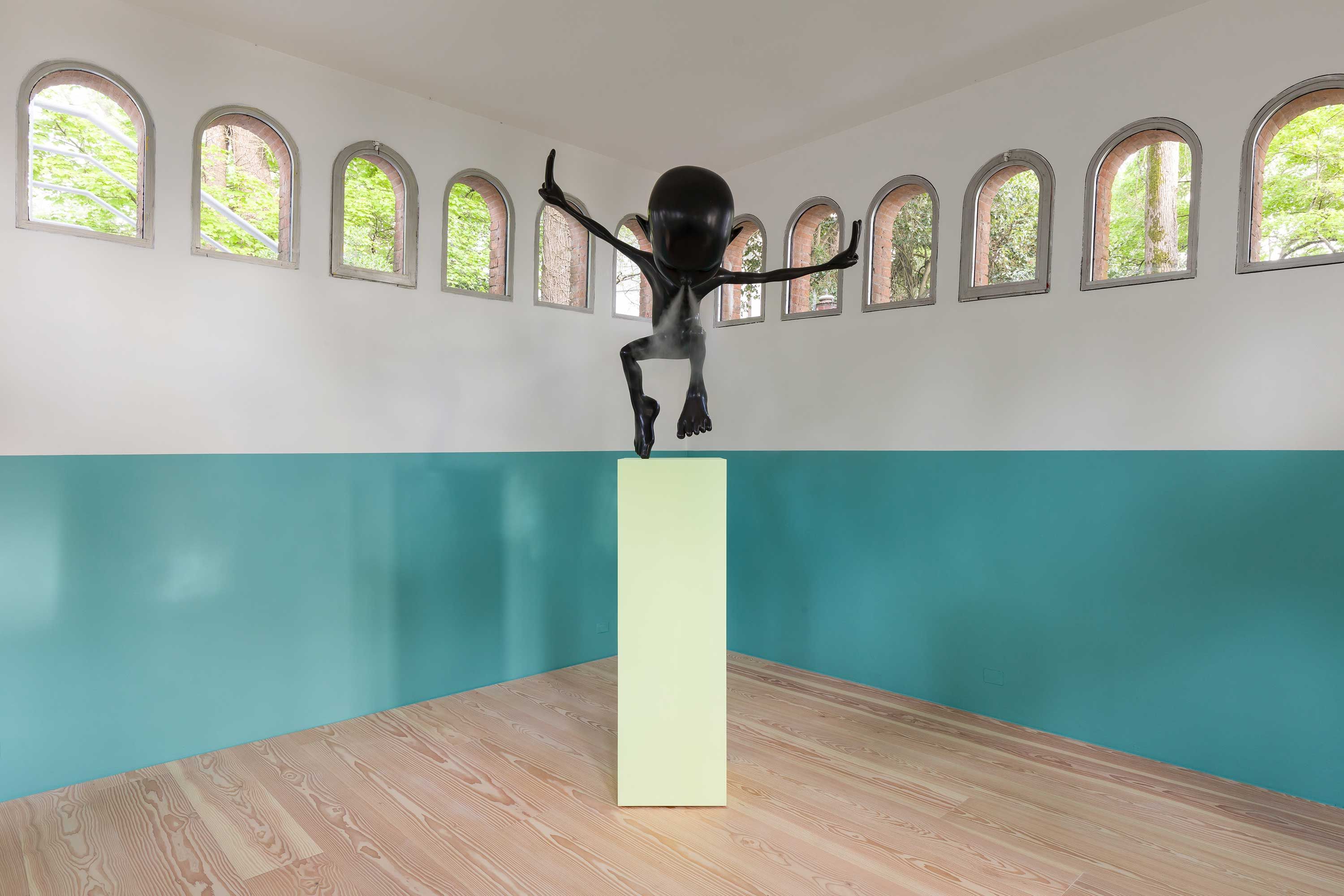 Art installation in a bright room with arched windows showing outside greenery. Features a glossy black humanoid sculpture...