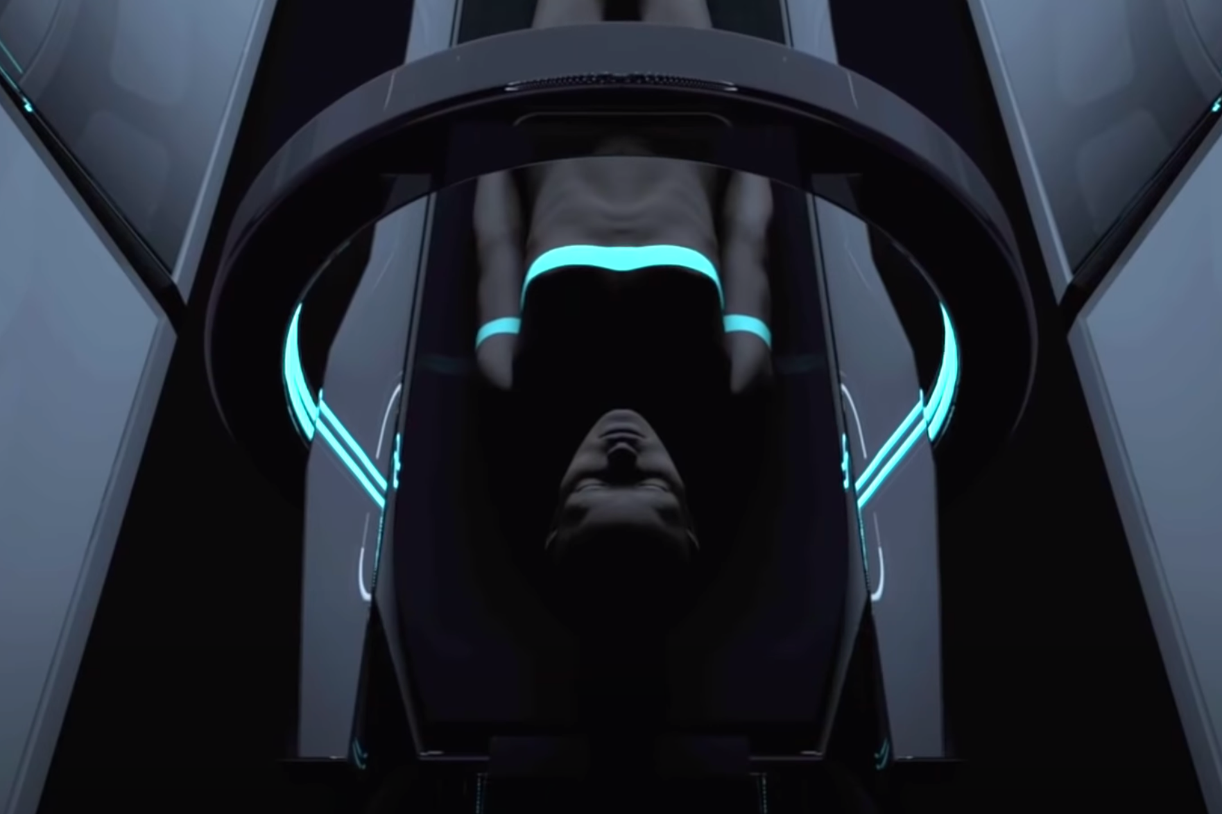 An individual is lying supine under an aqua blue scanner inside a dim and narrow room.