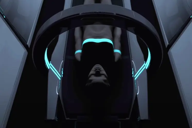 An individual is lying supine under an aqua blue scanner inside a dim and narrow room.