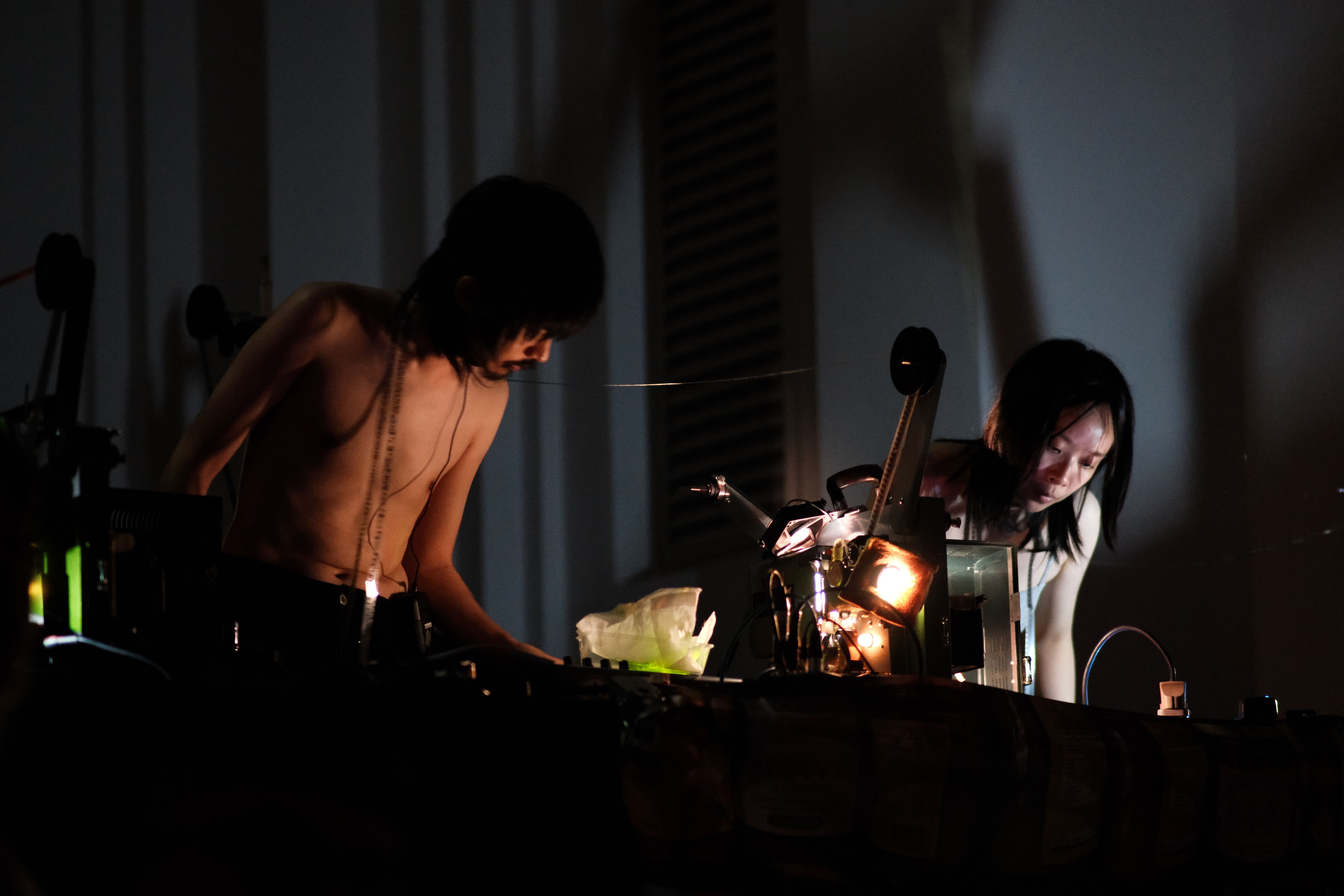 Two topless performers manipulate film projectors during an intense, low-light live performance.