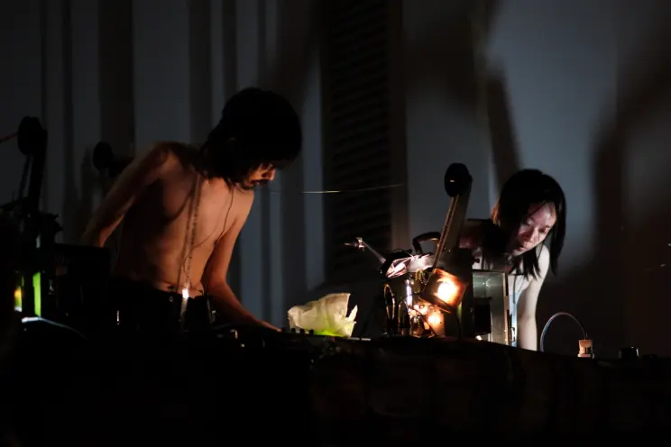 Two topless performers manipulate film projectors during an intense, low-light live performance.