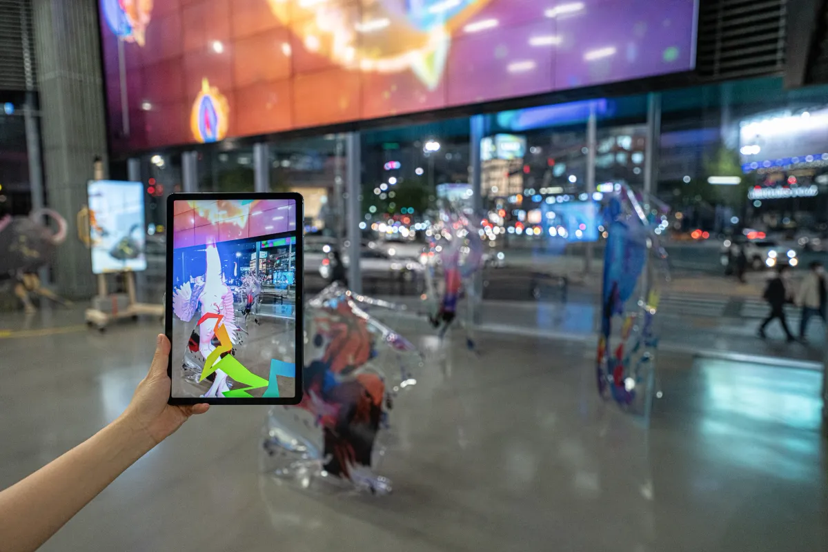 A hand holds a tablet displaying an art photo in sharp focus, contrasting with the blurry backdrop of a gallery hall.