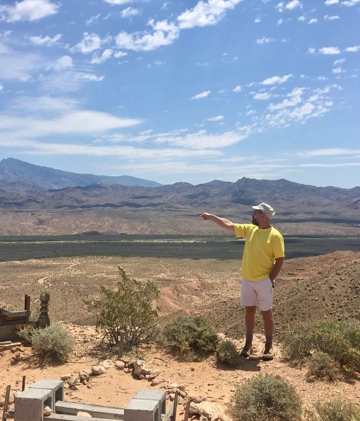 A person points into the distance while standing in a natural setting, with mountains, sky, and plants visible.