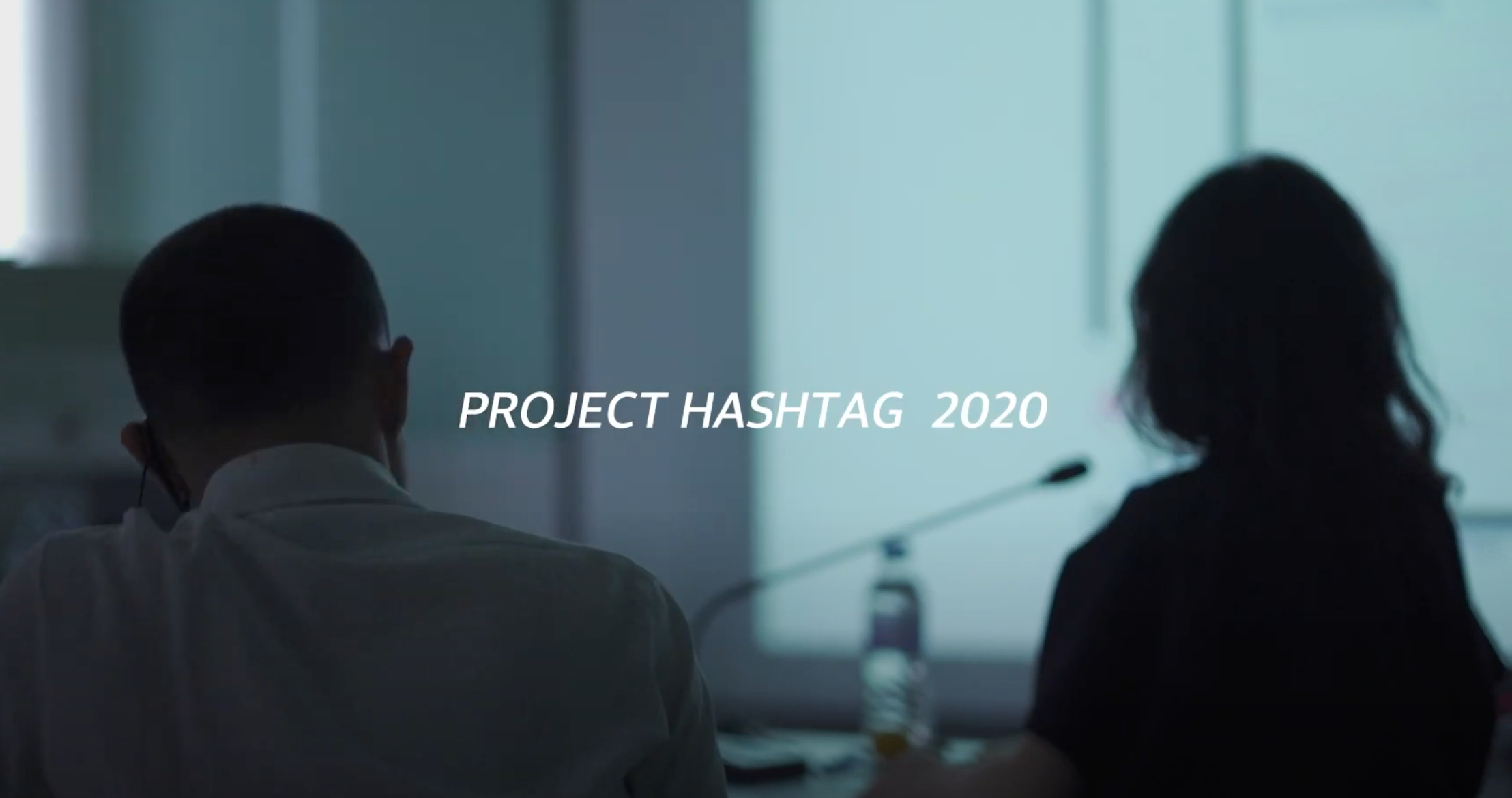 Two individuals engaged in a conference room meeting, viewed from behind. The text "Project Hashtag 2020" is prominently d...