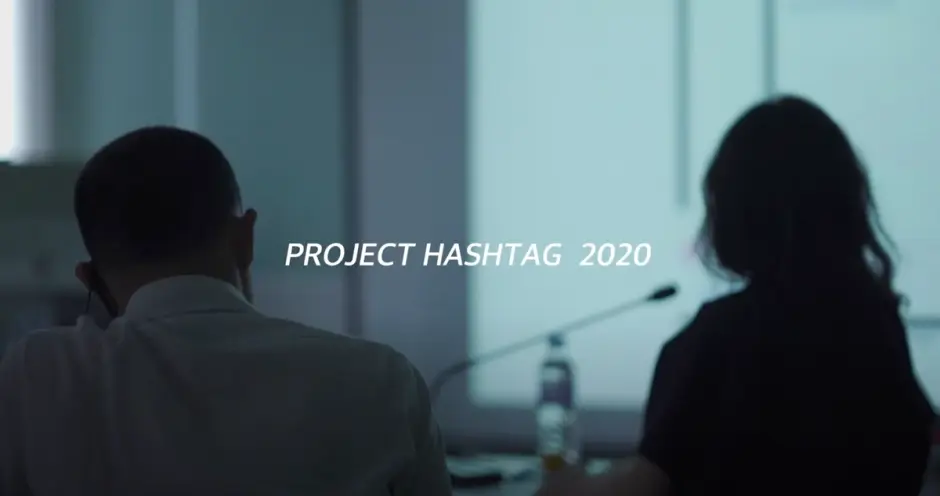 Two individuals engaged in a conference room meeting, viewed from behind. The text "Project Hashtag 2020" is prominently d...