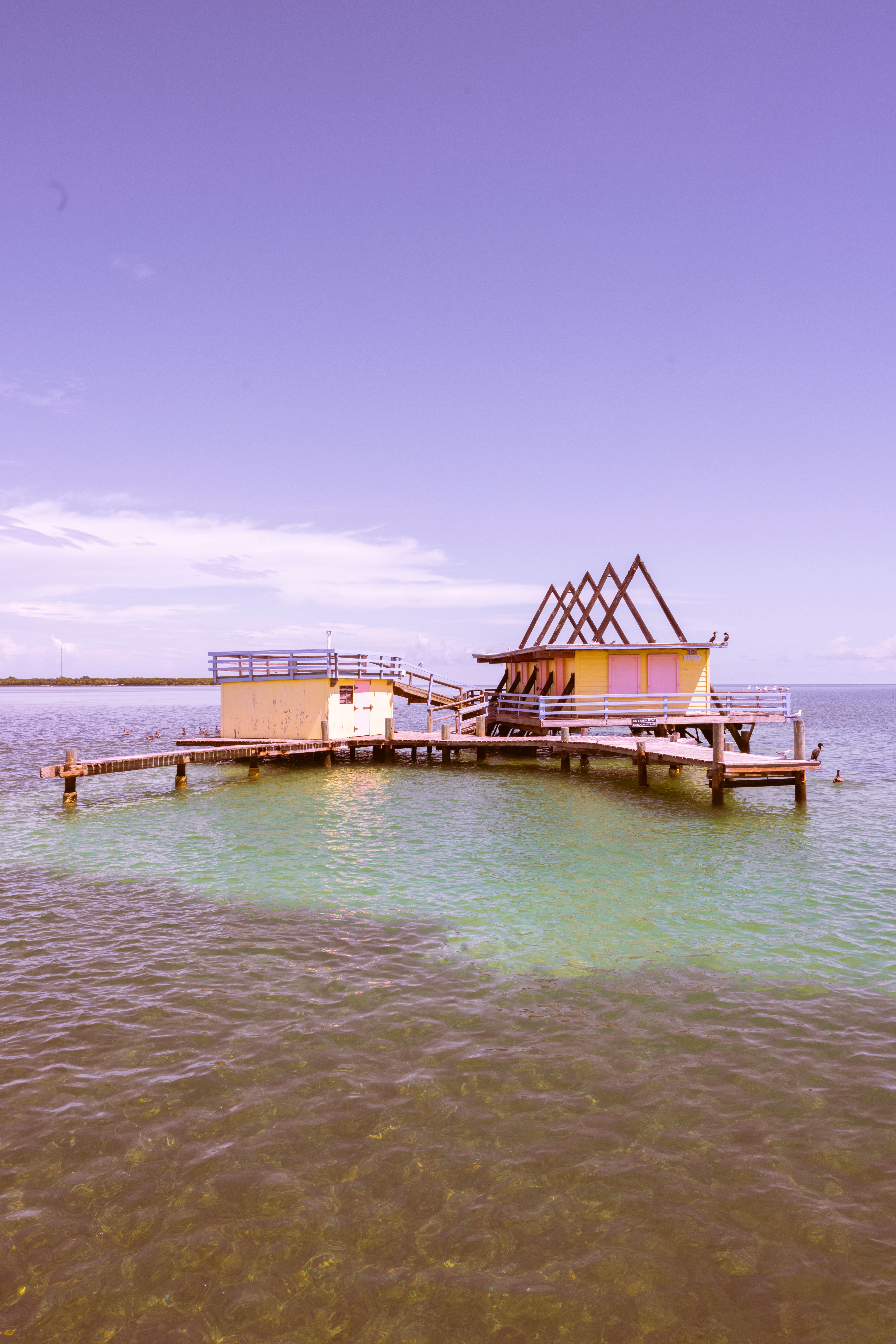 A pink-hued photo of a rustic wooden dock and hut over clear shallow waters.