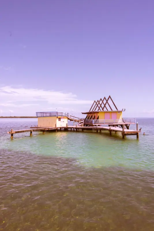 A pink-hued photo of a rustic wooden dock and hut over clear shallow waters.