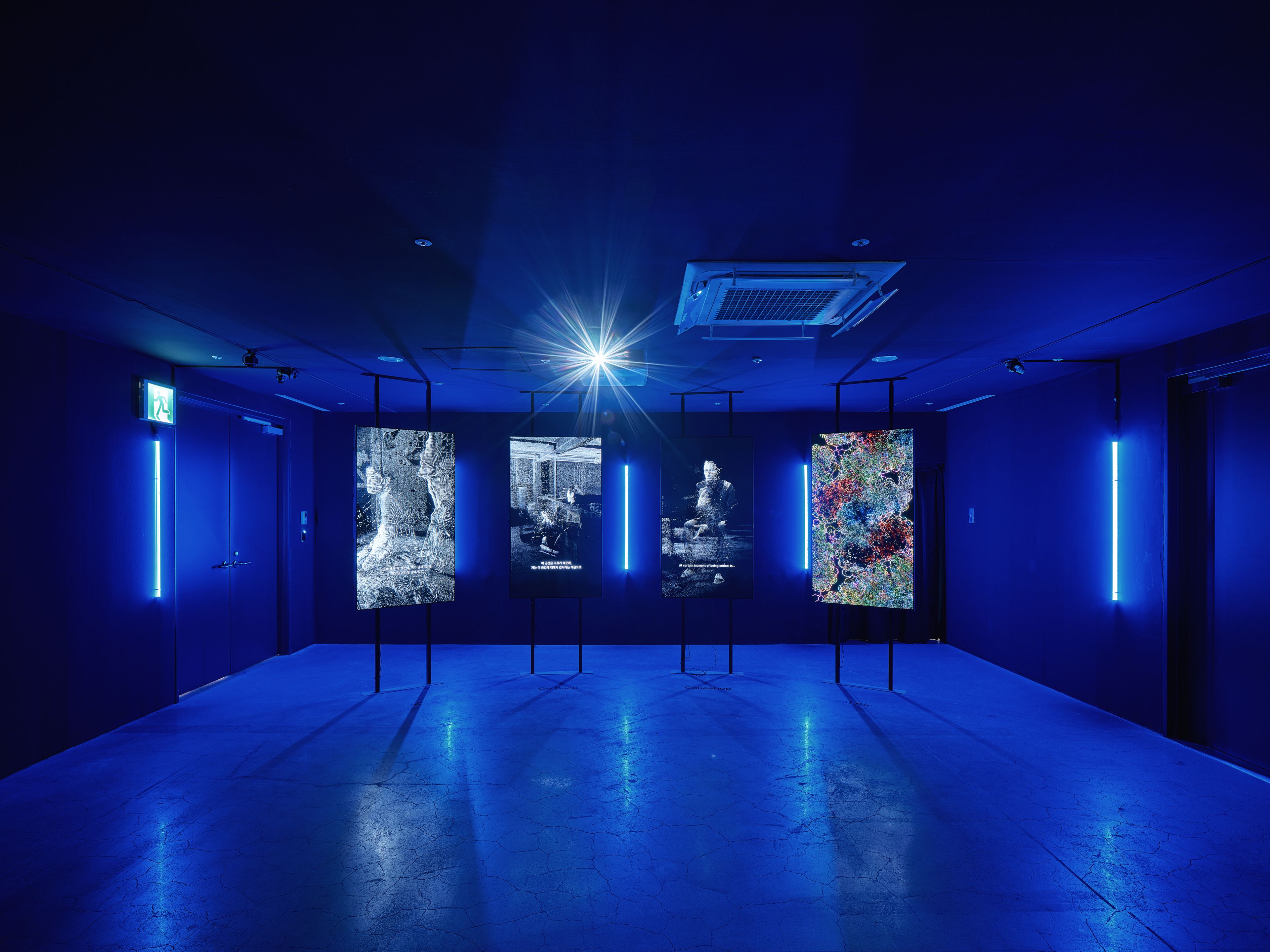 A dark room illuminated by soft blue lights houses an exhibition of four works: three monochrome posters and one colorful ...
