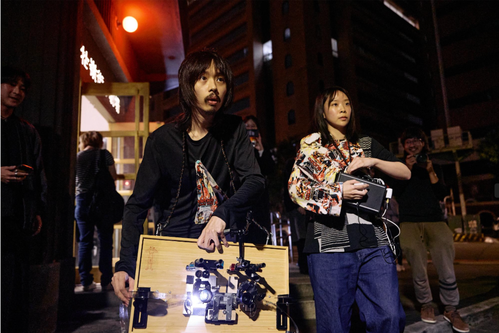 Two performers stand outside at night holding analog projection equipment, surrounded by onlookers.