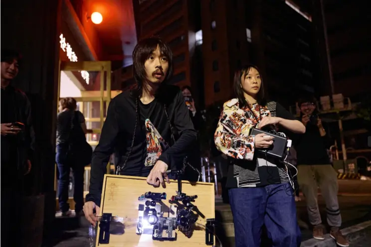 Two performers stand outside at night holding analog projection equipment, surrounded by onlookers.