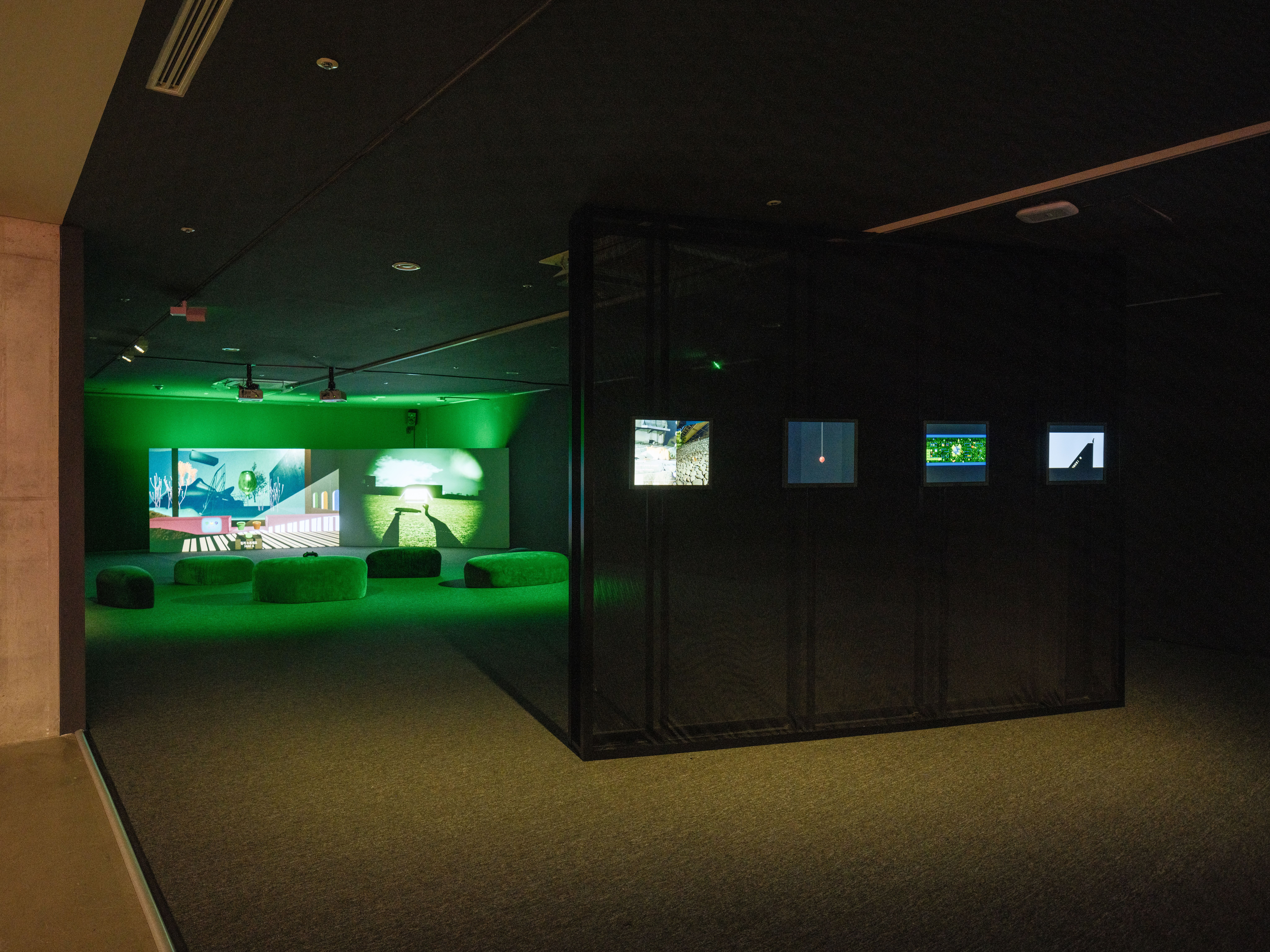 A dimly lit gallery cast in green light, with a wide projection screen at the far end displaying split-screen video footag...