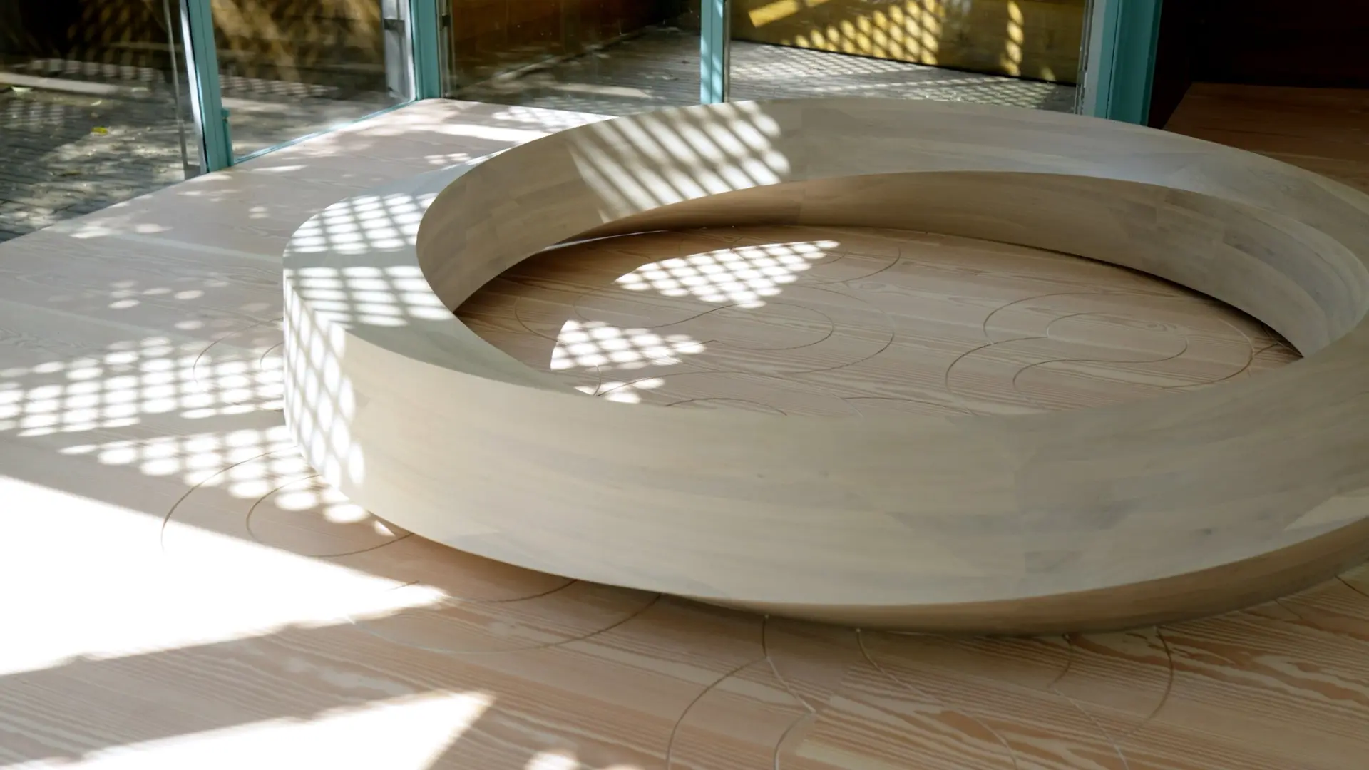 A circular wooden sculpture on a patterned wooden floor, with sunlight casting intricate shadows across the surface.
