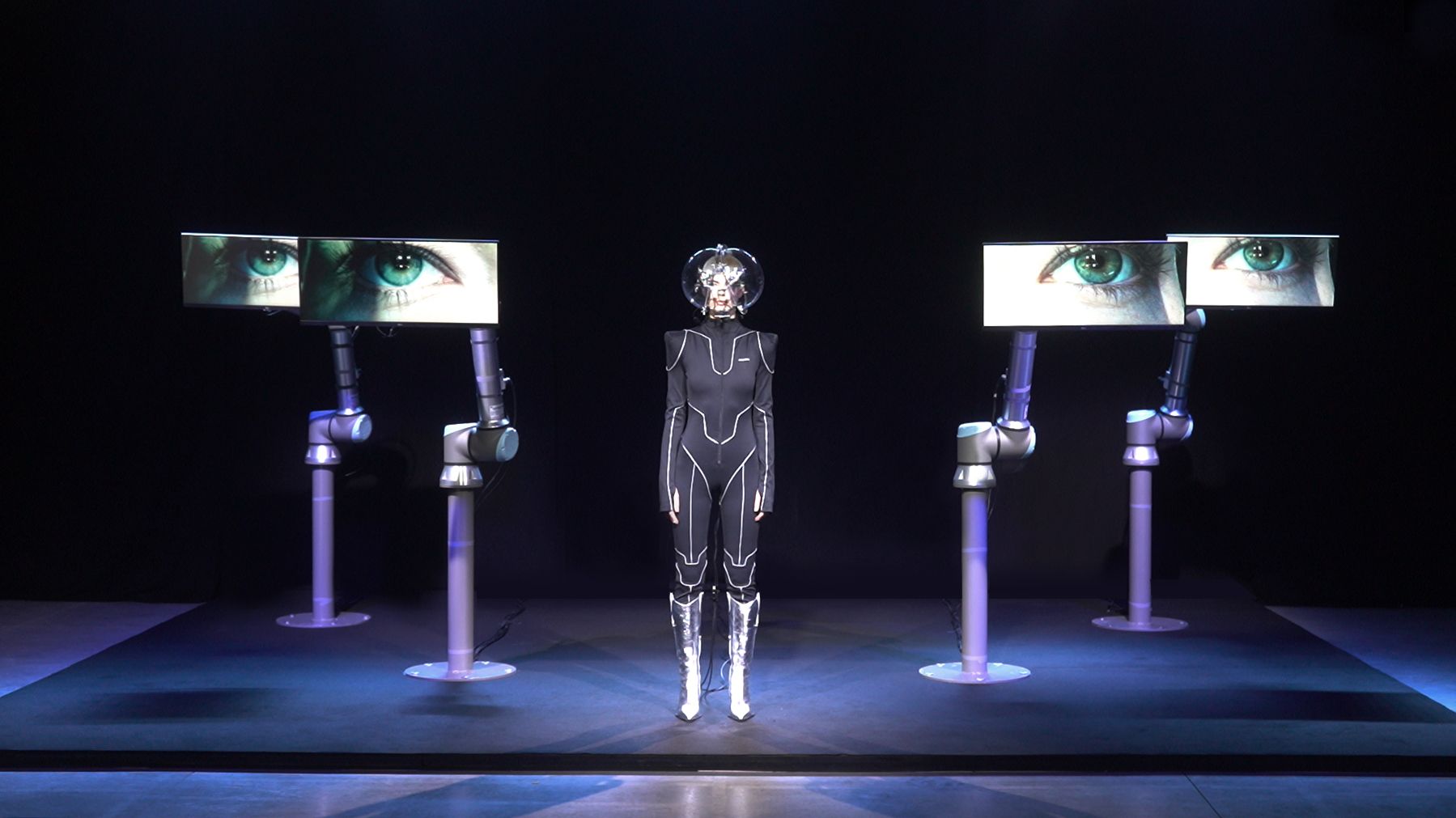 A person in a futuristic suit stands surrounded by robotic arms displaying close-up images of eyes on monitors in a dark s...