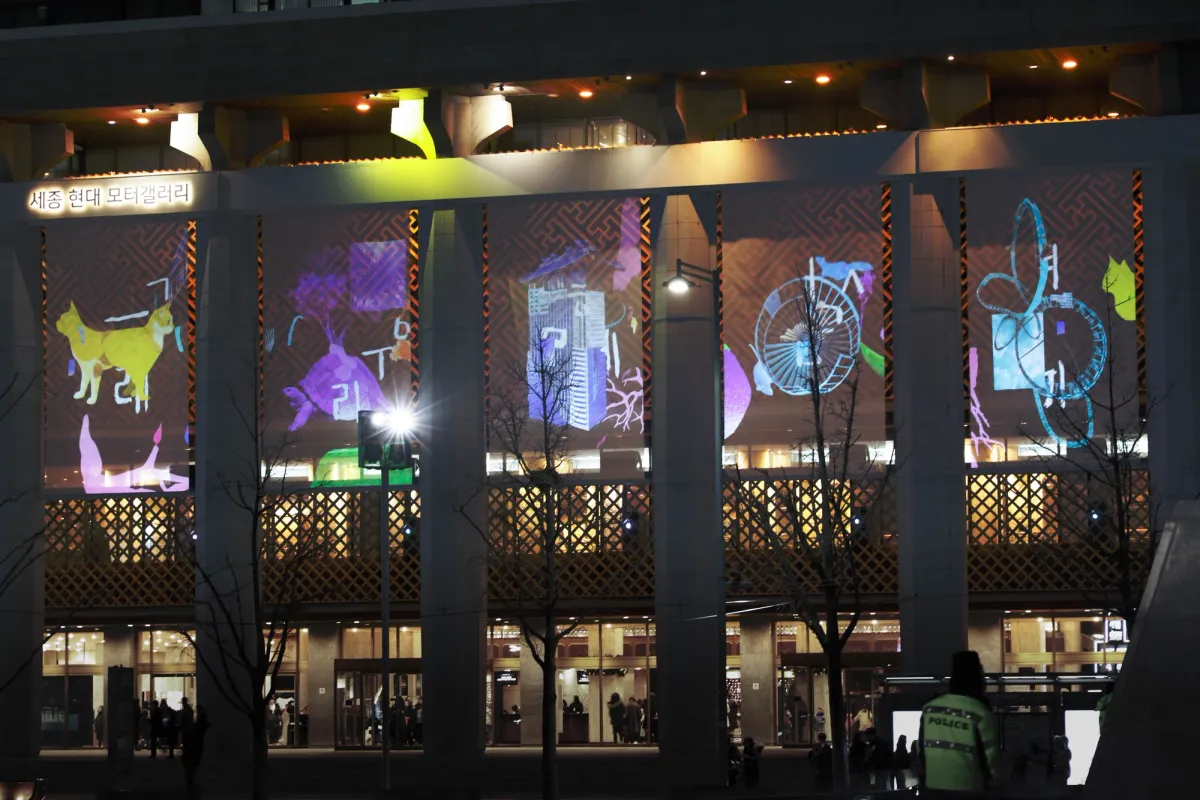 The façade of a building is transformed into a vibrant canvas, with projected images of various objects including a cat, a piano, and a fan creating a stylized visual montage.