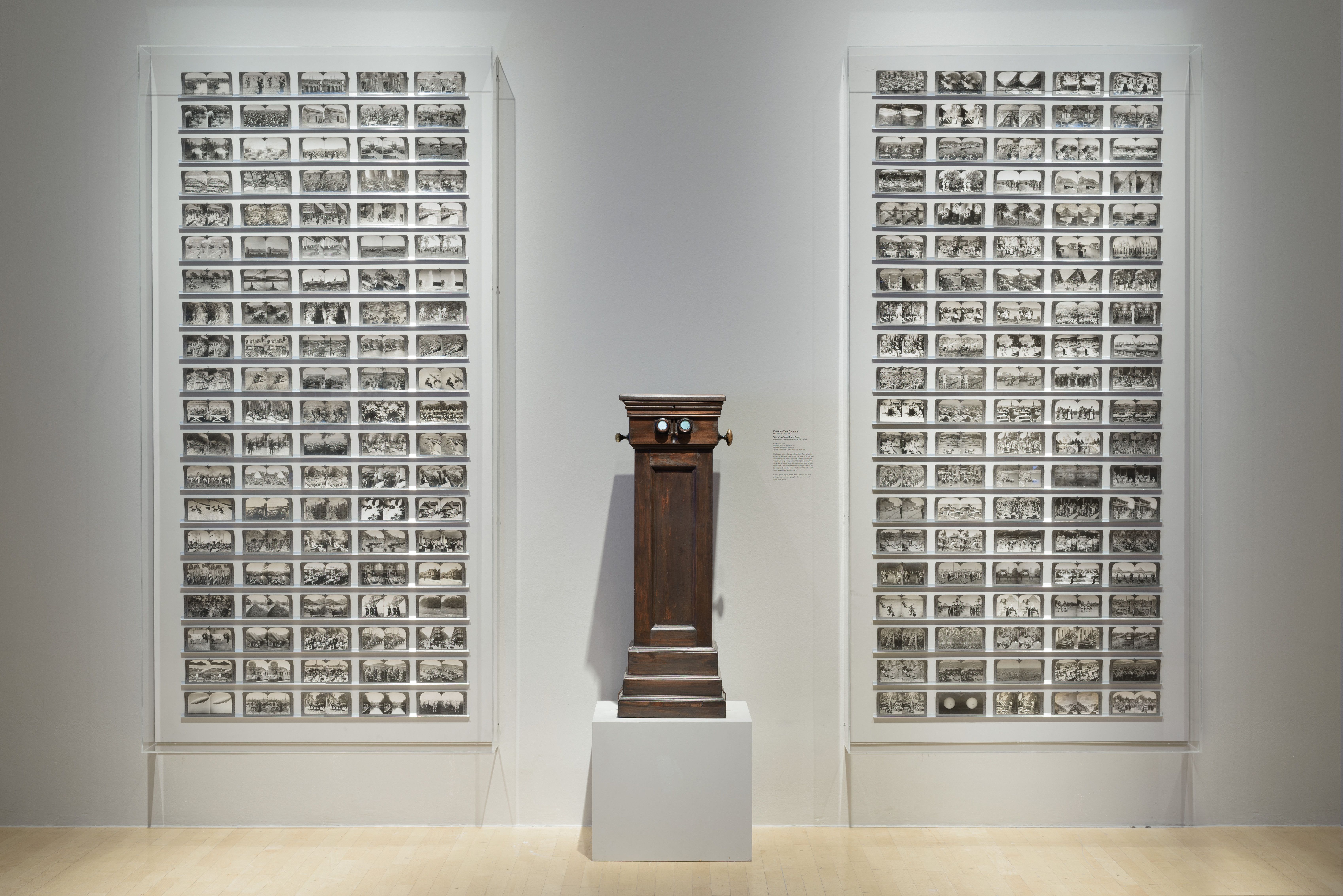 Two large frames filled with black and white photographs arranged in five columns hang on a white wall. Situated between t...