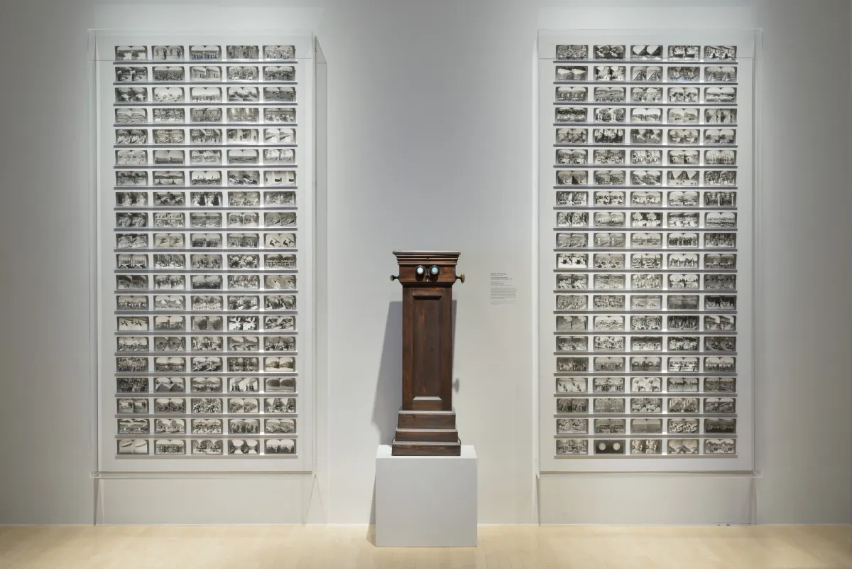 Two large frames filled with black and white photographs arranged in five columns hang on a white wall. Situated between them on a white pedestal is a vintage wooden stereoscope viewer.