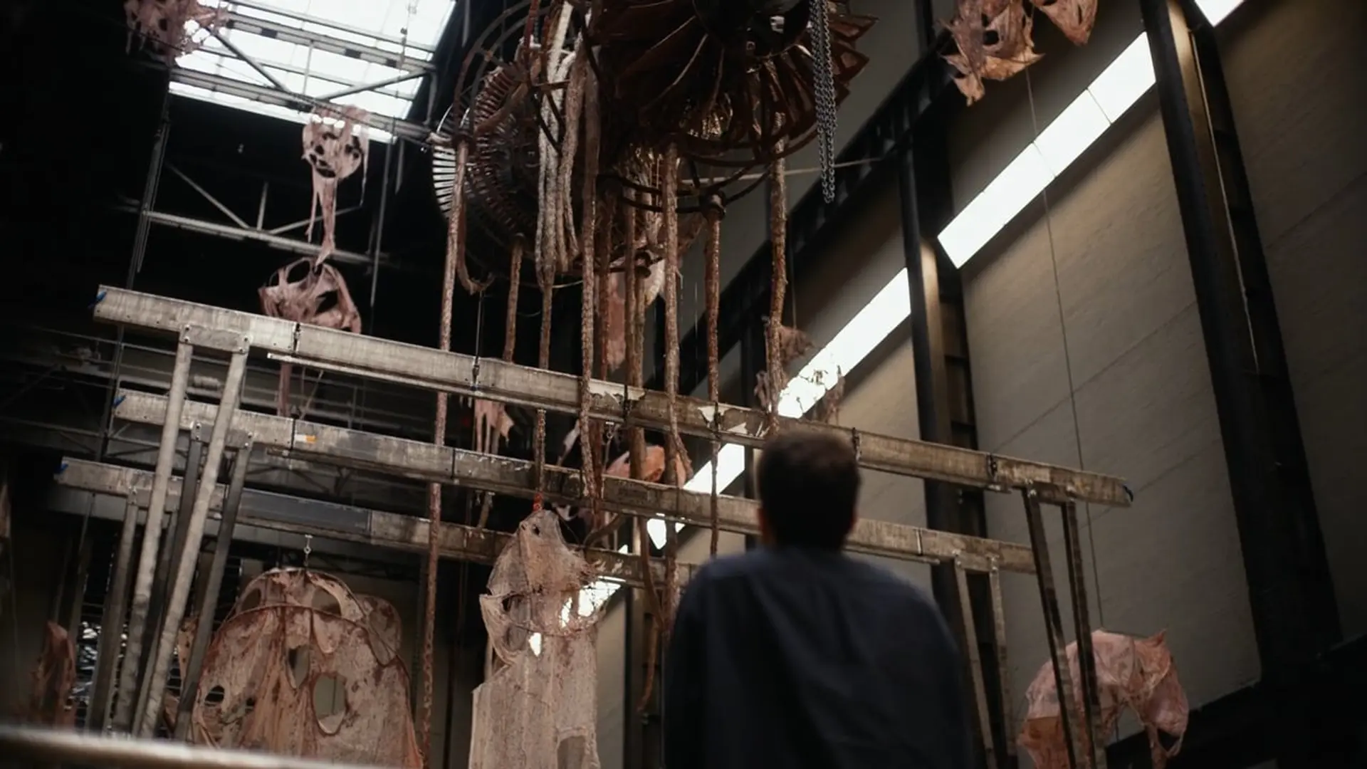Person observing large, industrial art installation with suspended, weathered metal and fabric elements.