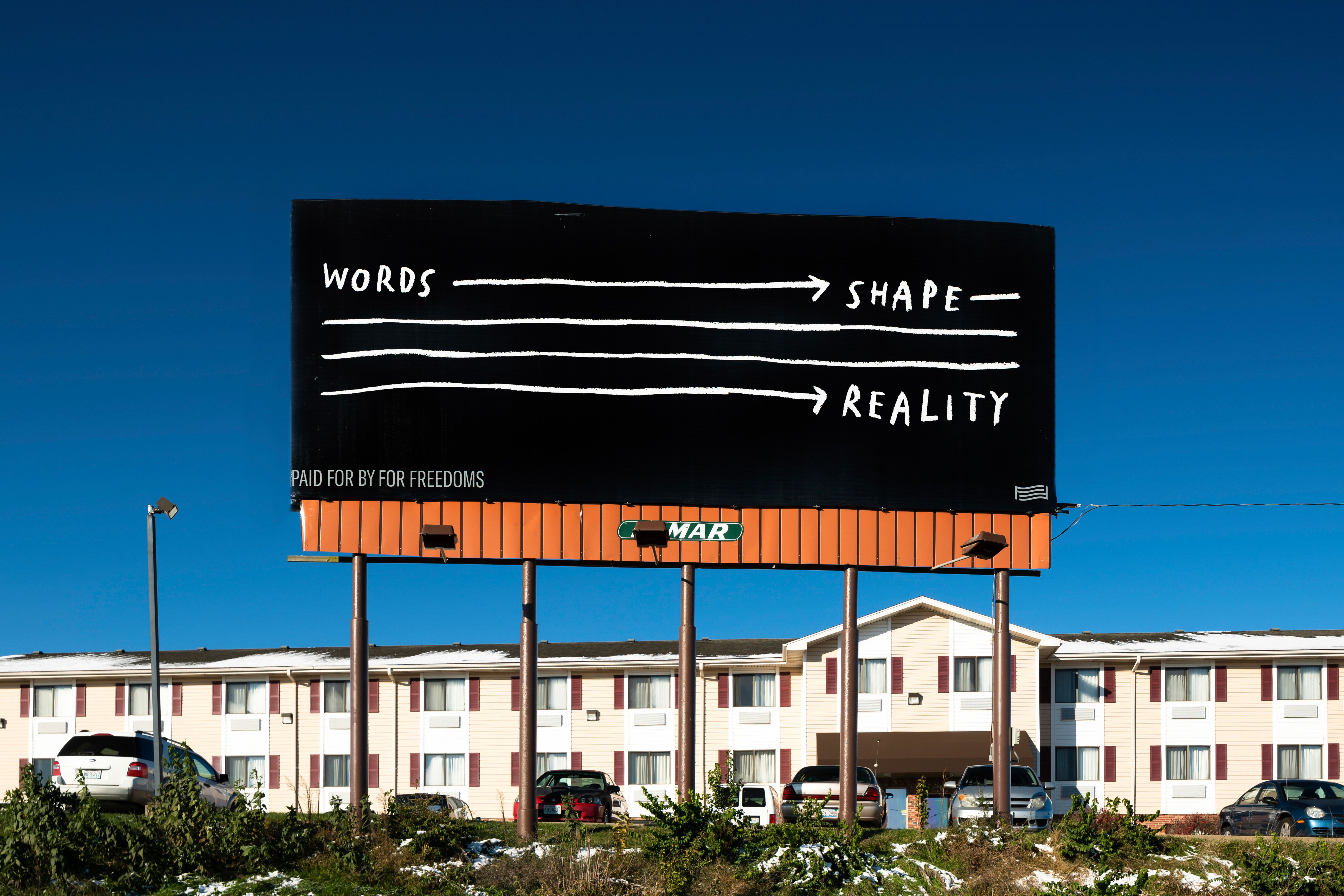 A roadside billboard reading “WORDS SHAPE REALITY” in white text on a black background.