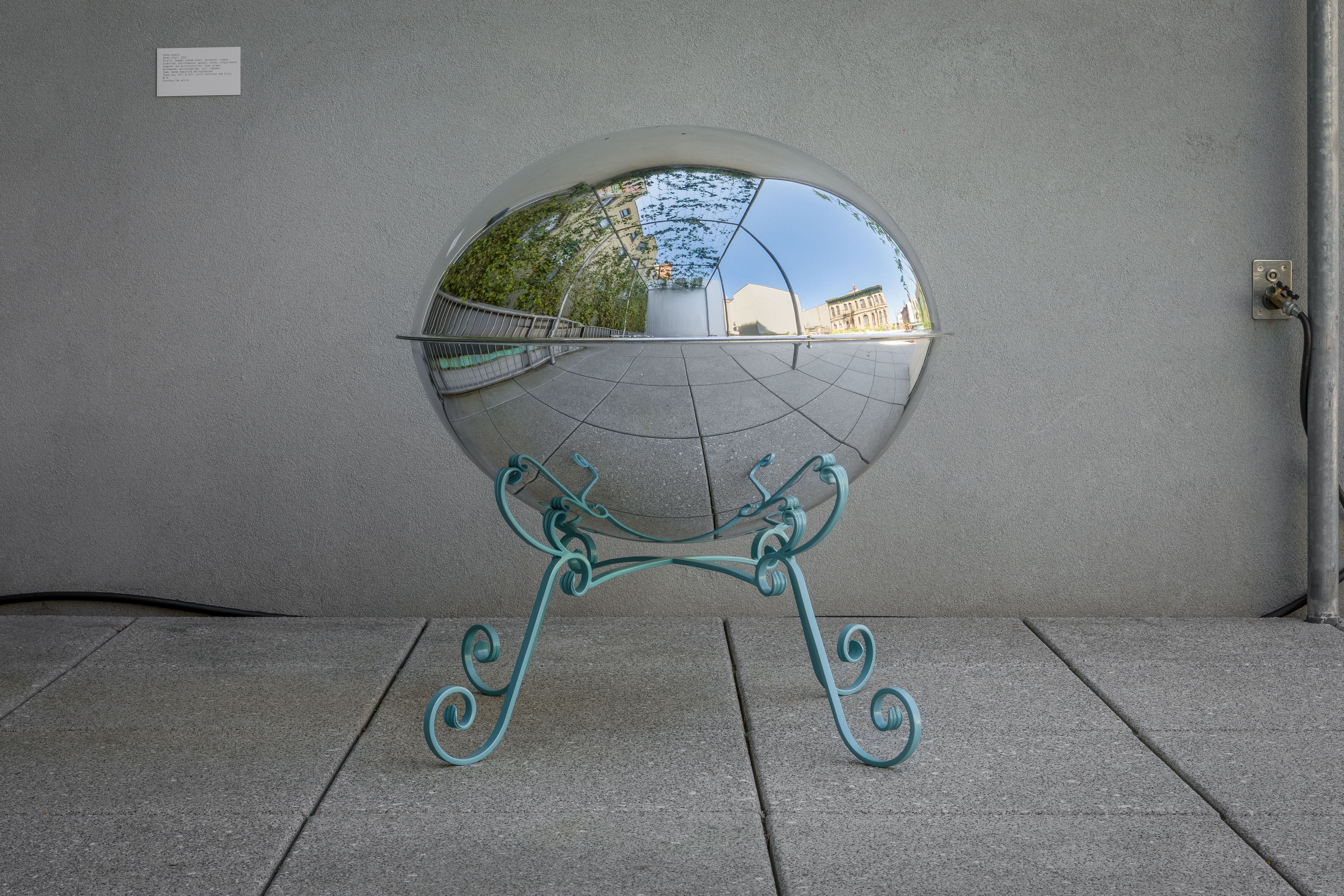 A reflective hemispherical sculpture on ornate teal legs, mirroring an outdoor terrace and buildings.