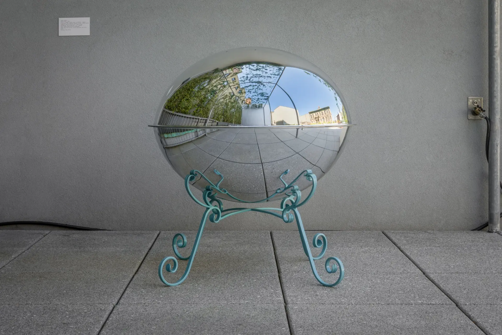 A reflective hemispherical sculpture on ornate teal legs, mirroring an outdoor terrace and buildings.