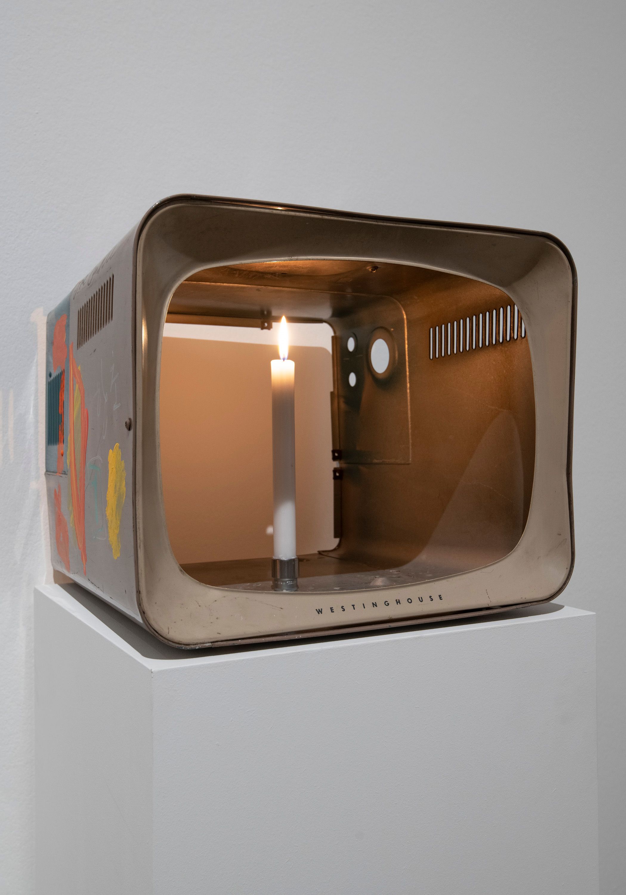 An old Westinghouse portable TV, coated with paint on its sides, houses a lit candle within. It's showcased against a whit...