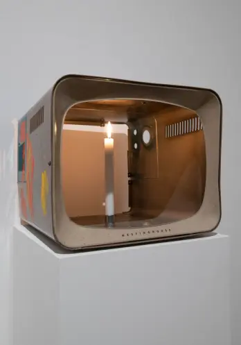 An old Westinghouse portable TV, coated with paint on its sides, houses a lit candle within. It's showcased against a whit...