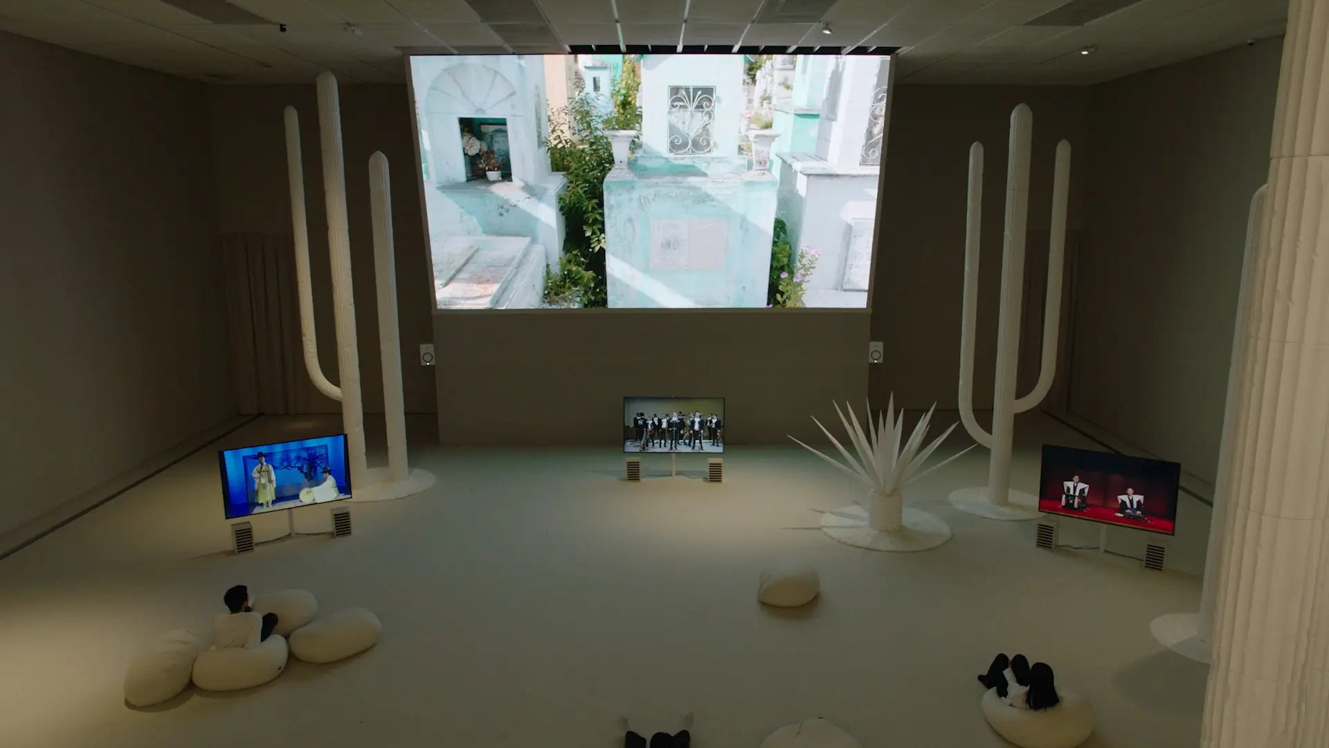 Gallery room with video art displays, white sculptures resembling cacti, and visitors on beanbag seats.