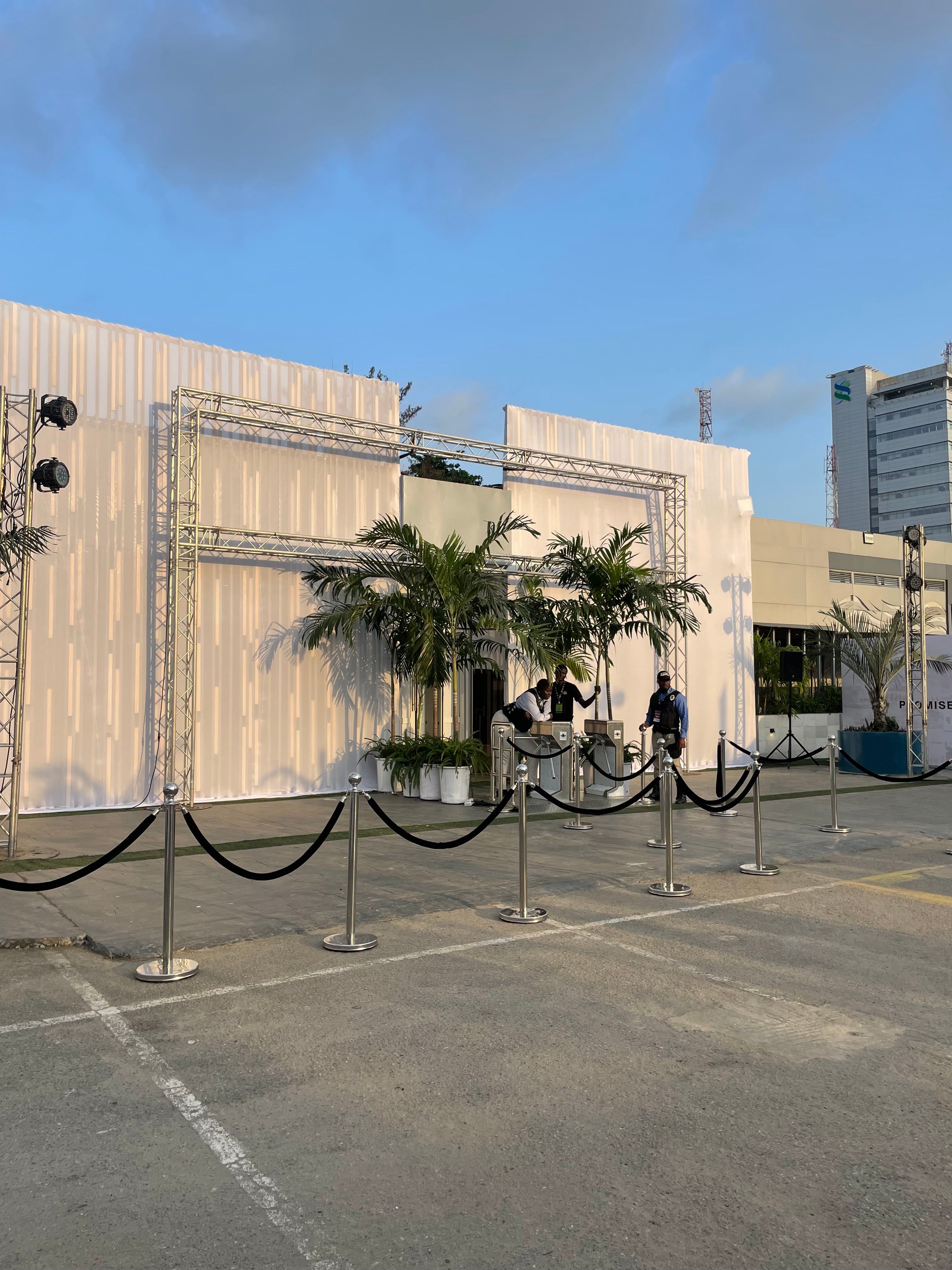 An elegant outdoor event setup with white drapery, palm plants, and black velvet ropes.