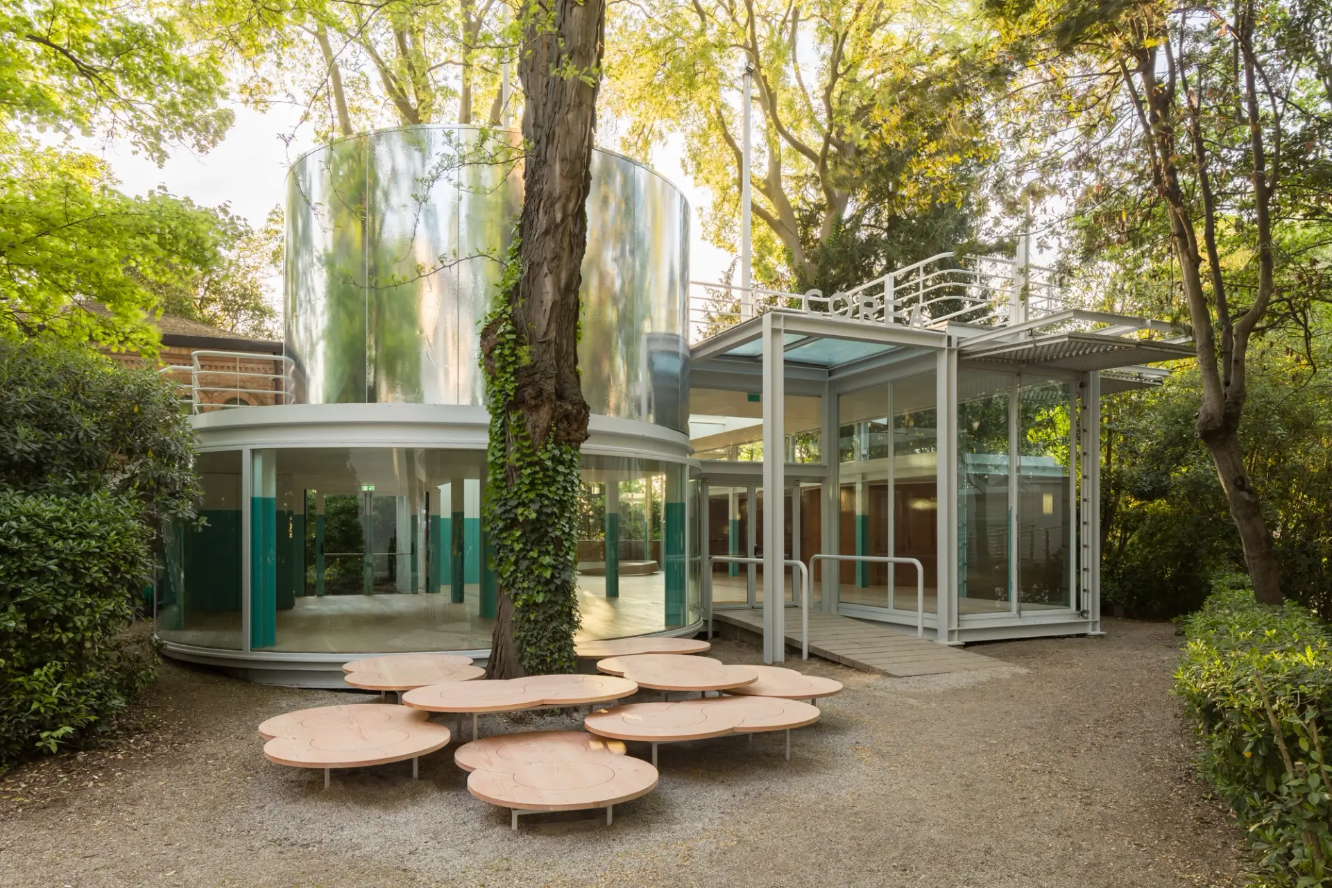 Mirrored cylindrical structures, glass walls, and round seating in a wooded outdoor installation at the Korean Pavilion 2024.