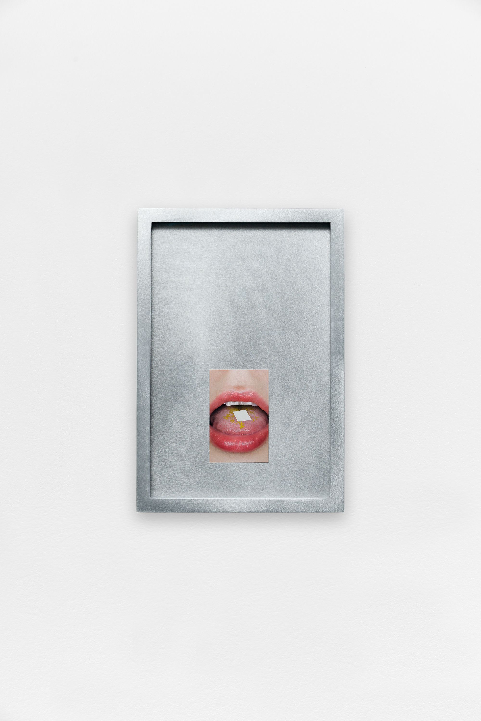 A small photograph of a mouth with a tab on the tongue, centered within a brushed metal frame on a white wall.