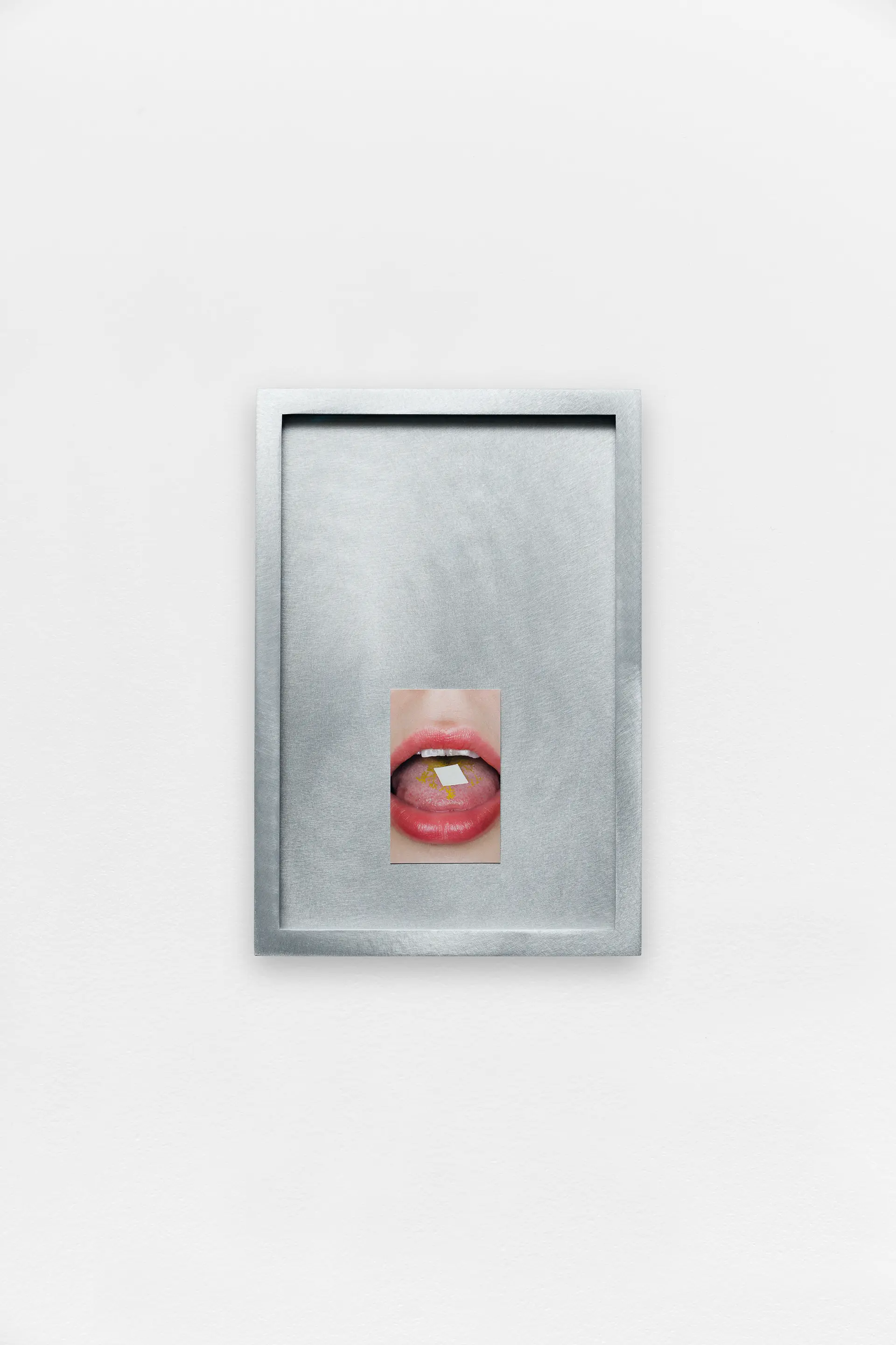 A small photograph of a mouth with a tab on the tongue, centered within a brushed metal frame on a white wall.
