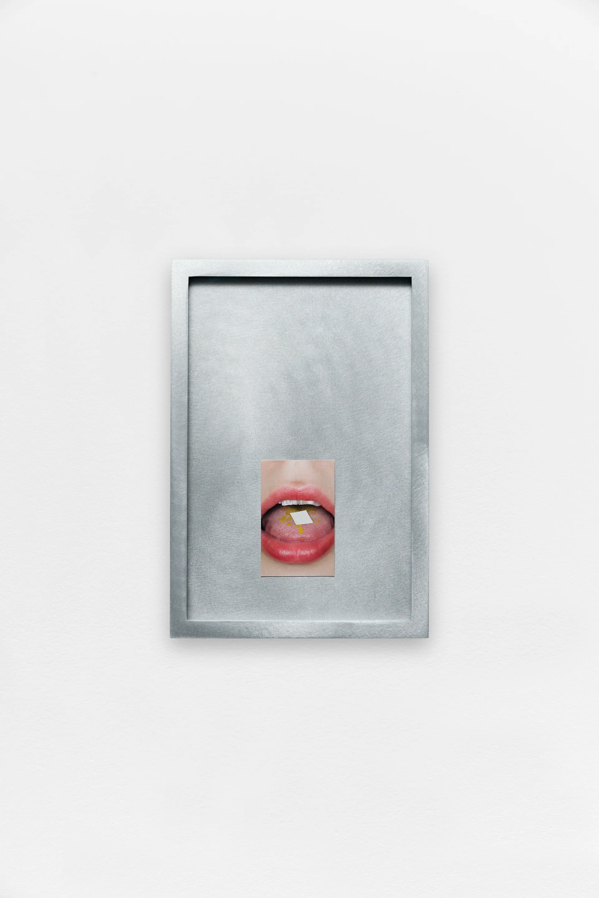A small photograph of a mouth with a tab on the tongue, centered within a brushed metal frame on a white wall.