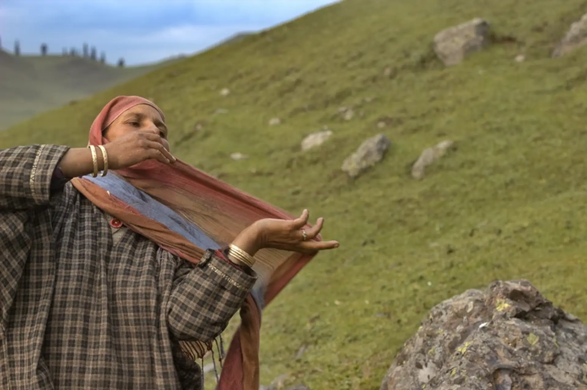 Woman adjusting shawl on a hill, green landscape in the background.