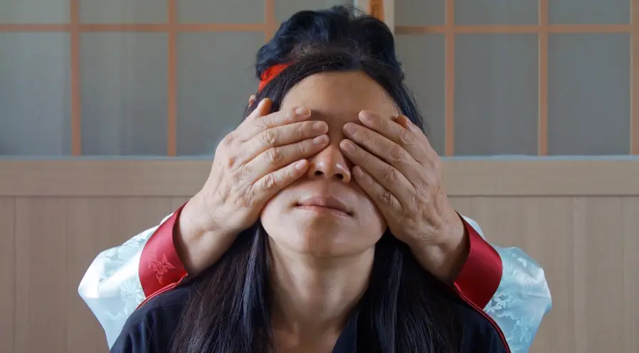A person stands in a still shot from the projected film "Community of Parting", 2019. Another person behind them covers th...