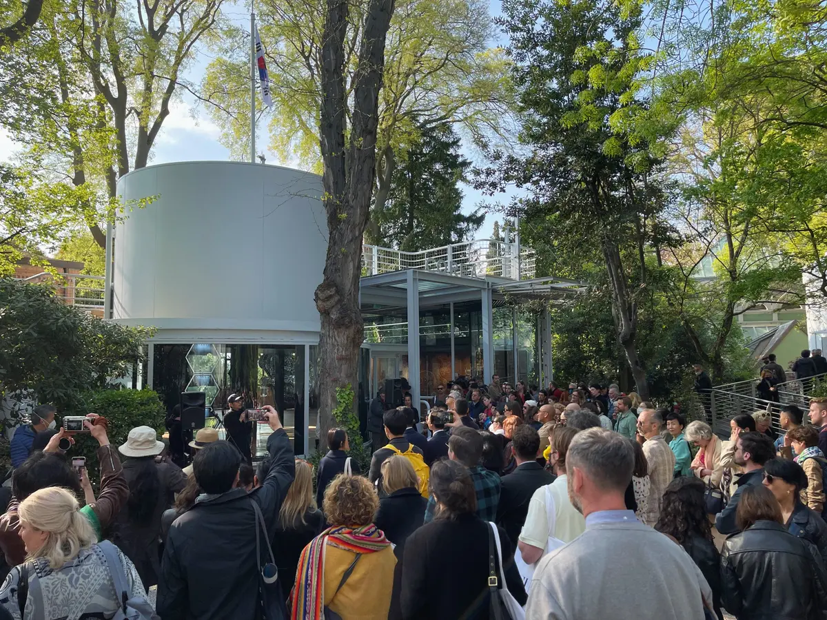 A crowd gathers in daylight outside an exhibition space, nestled amidst trees.