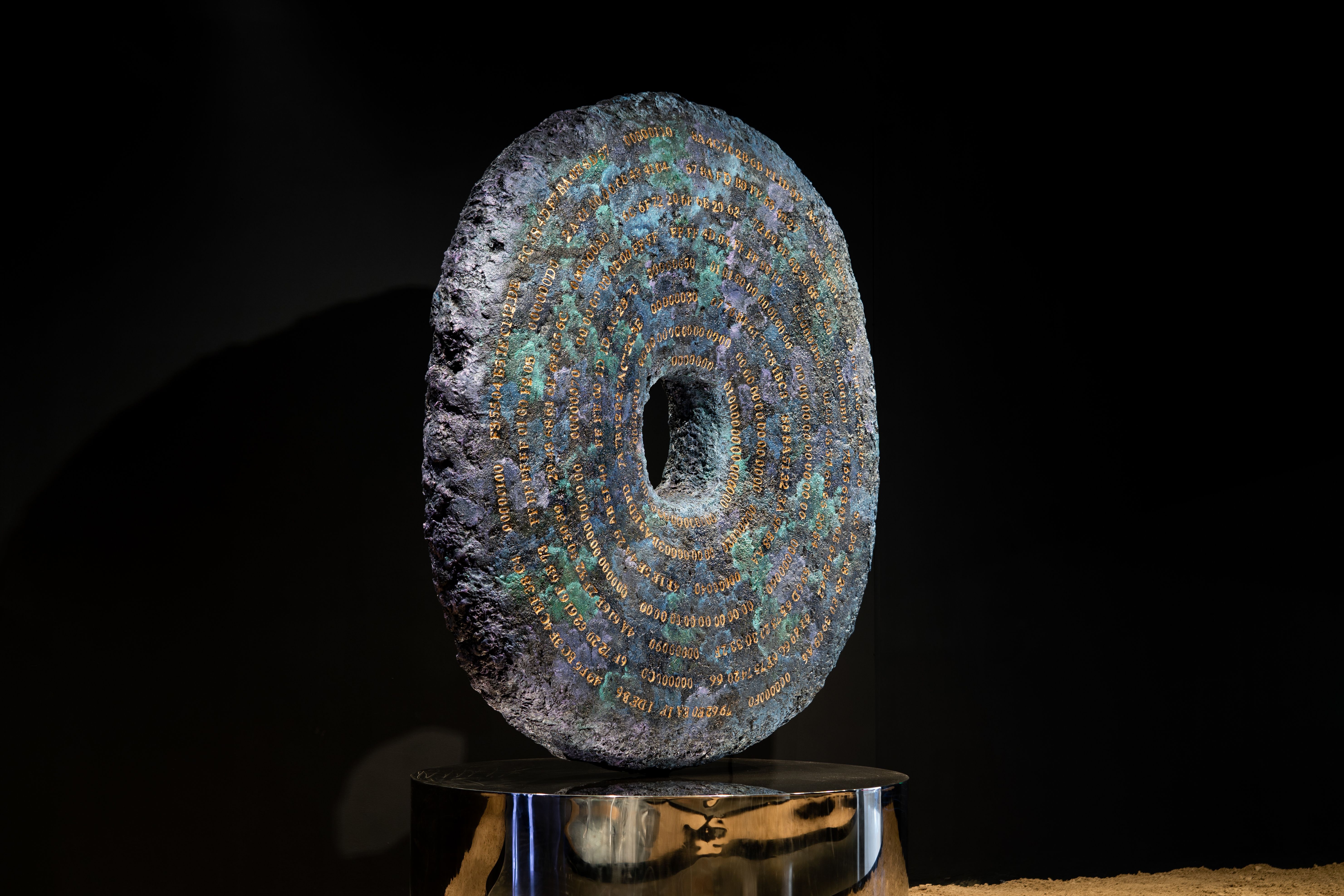 Dark circular textured sculpture with intricate blue details on shiny pedestal.