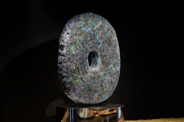 Dark circular textured sculpture with intricate blue details on shiny pedestal.