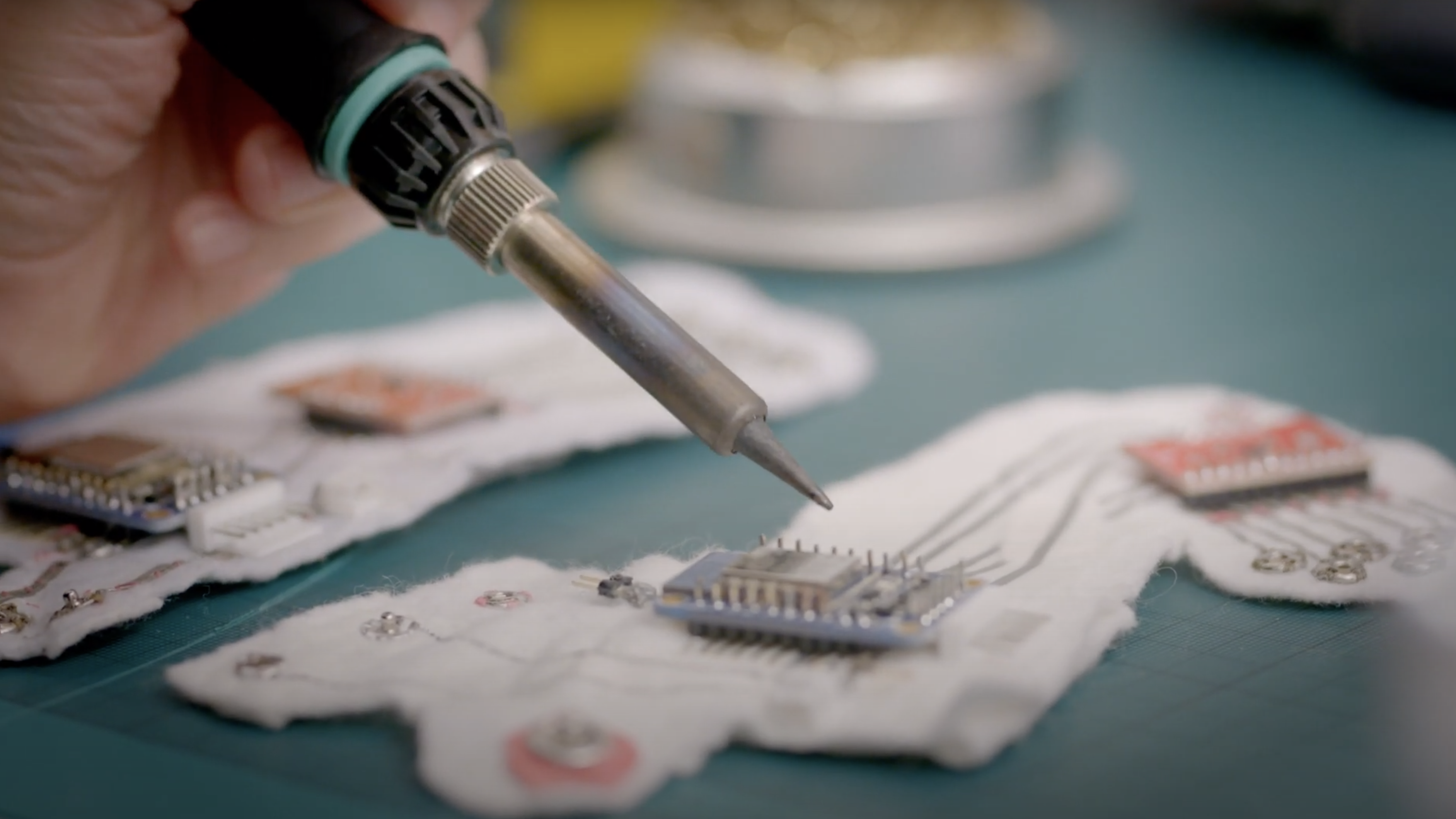 A close-up view of a hand using a tool to work on a microchip. The work is being conducted on a vibrant green surface.