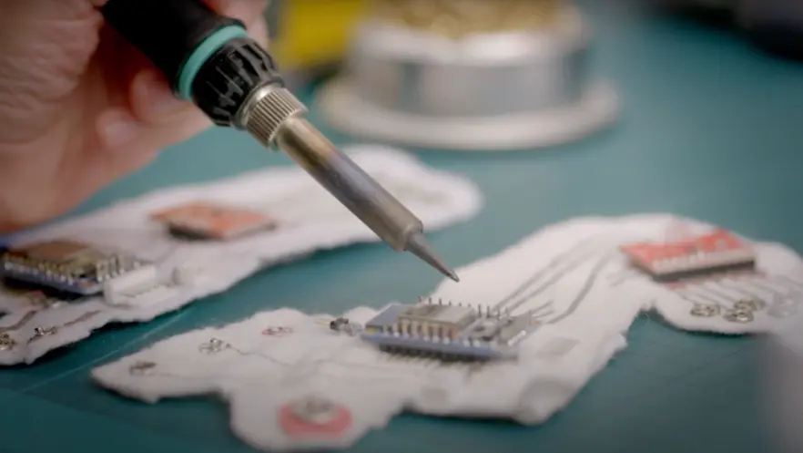 A close-up view of a hand using a tool to work on a microchip. The work is being conducted on a vibrant green surface.