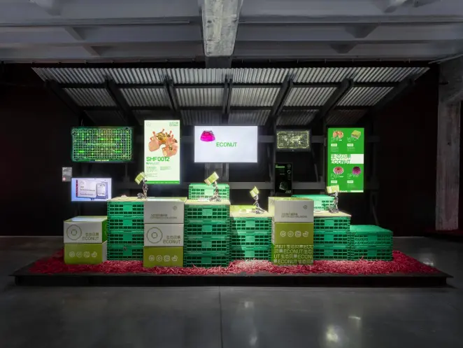 Exhibition installation with stacked green crates, screens displaying "ECONUT" and other graphics.