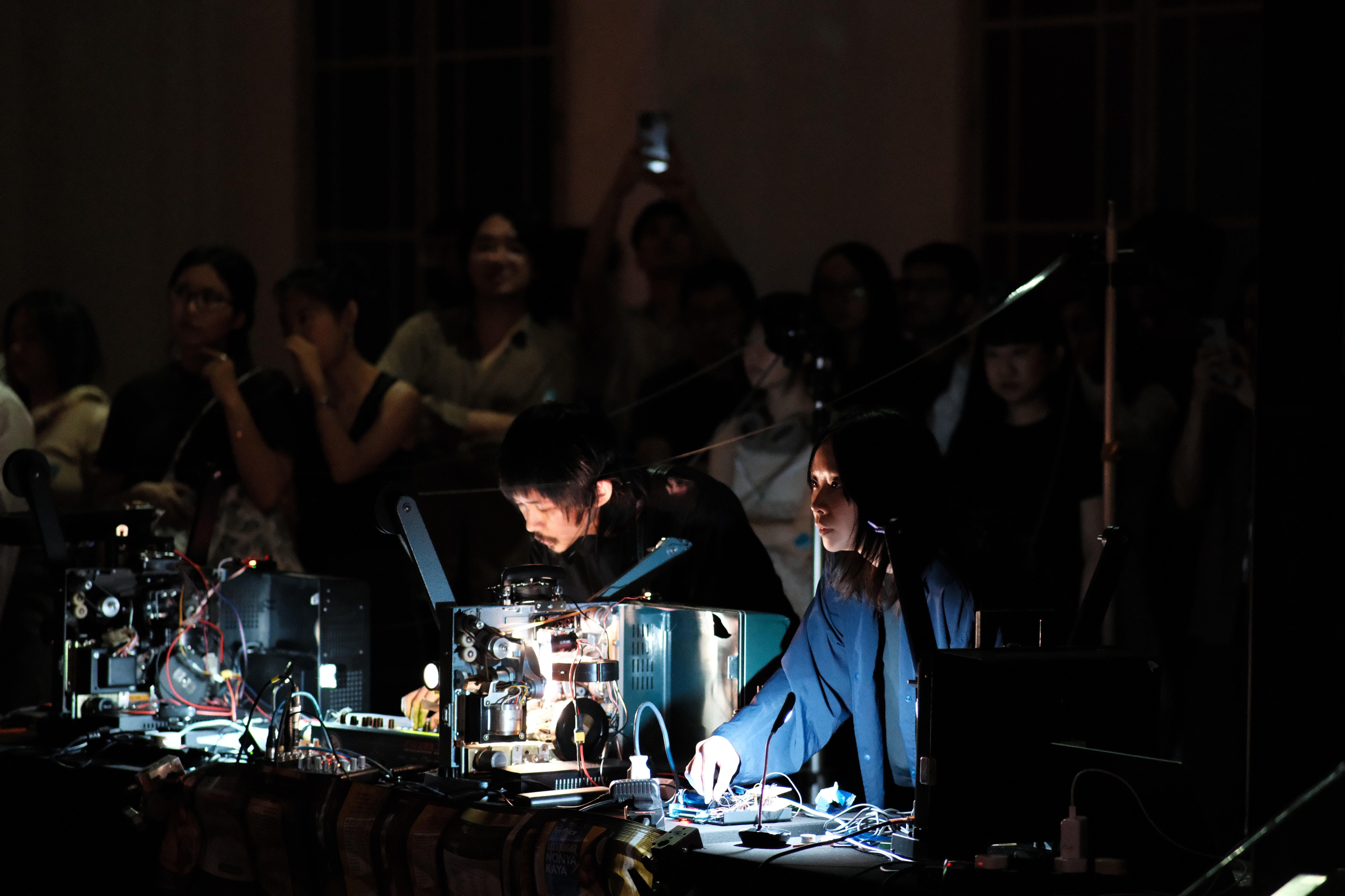 Two artists perform with illuminated projectors before a focused audience in a darkened space.