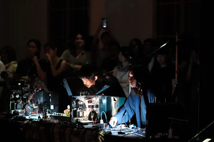 Two artists perform with illuminated projectors before a focused audience in a darkened space.