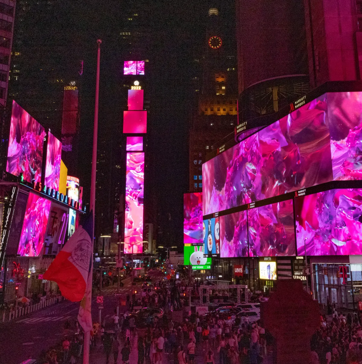 An abstract piece of pink arts is displayed on the advertisement screens of Times Square, surrounded by towering buildings. The vibrant artwork provides a stark contrast to the urban surroundings.