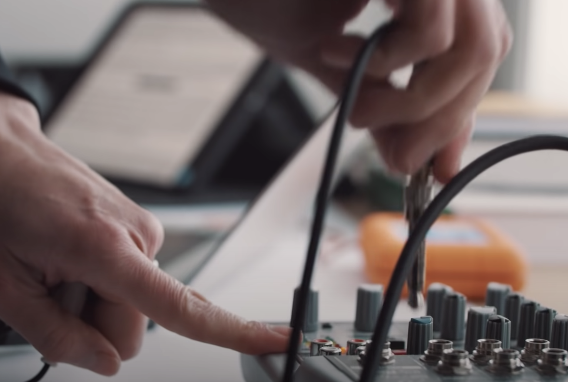 A pair of hands connecting a device to an array of audio equipment, cables visible. In the blurred background, a laptop sc...
