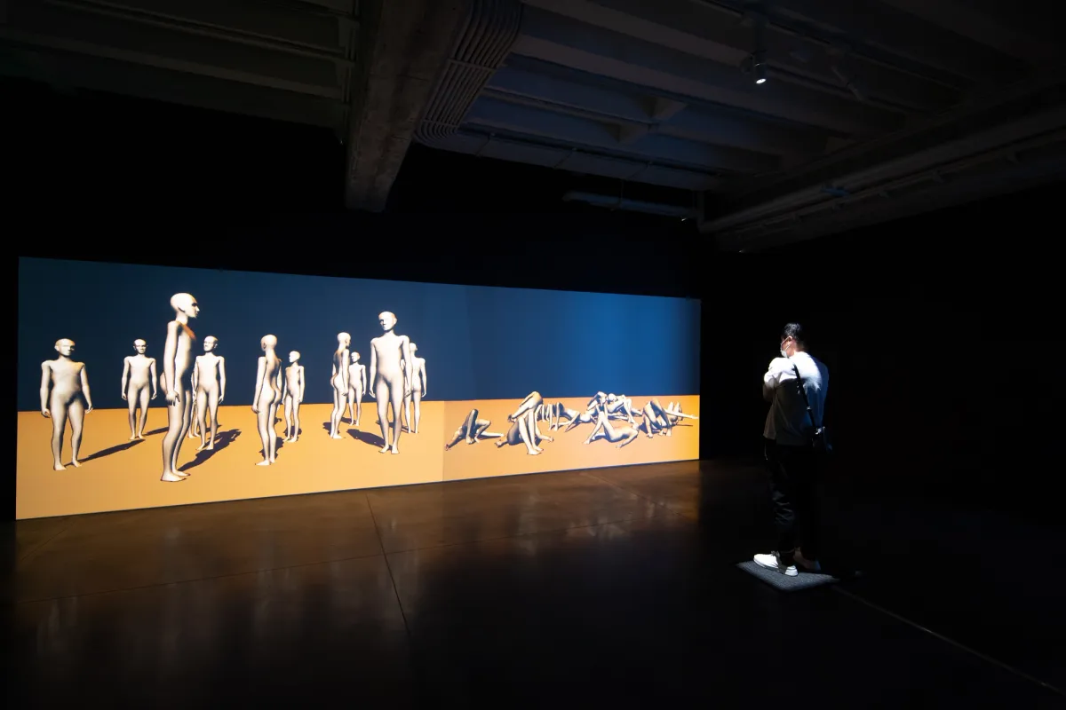 An expansive art piece depicting standing white human forms on the left side and reclining ones on the right, set against a two-toned background - yellow on the bottom and blue at the top, in a dim room.