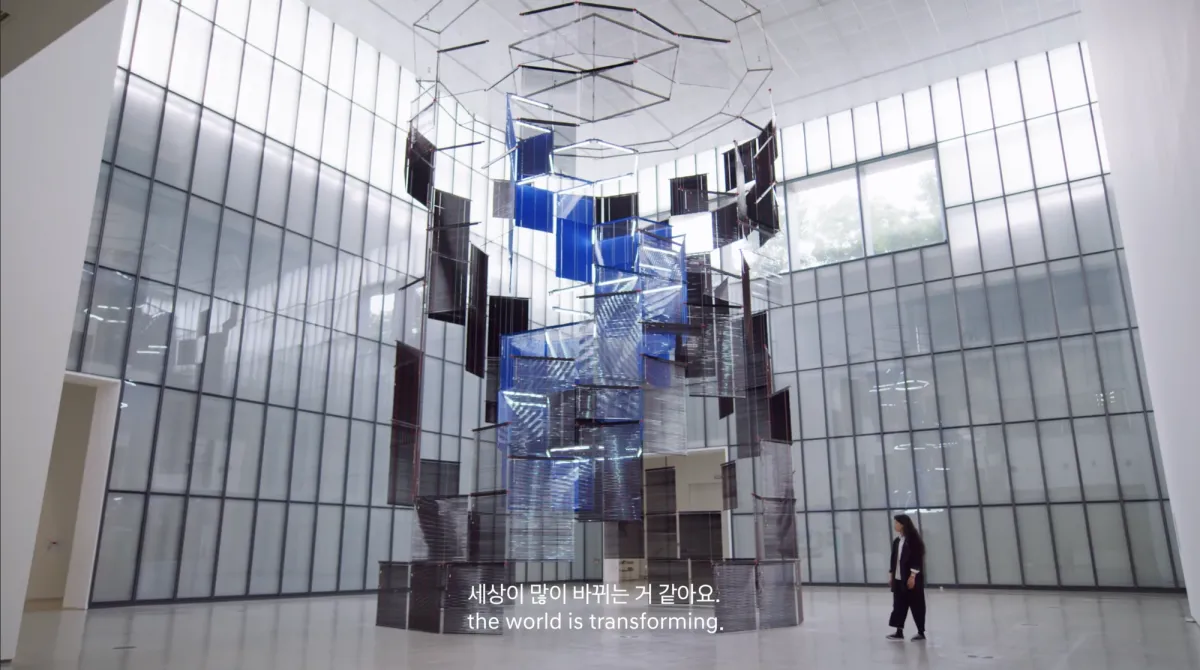 A visitor studying a blue and transparent panel art installation with text "세상이많이바뀌는거같아요. the world is transforming."
